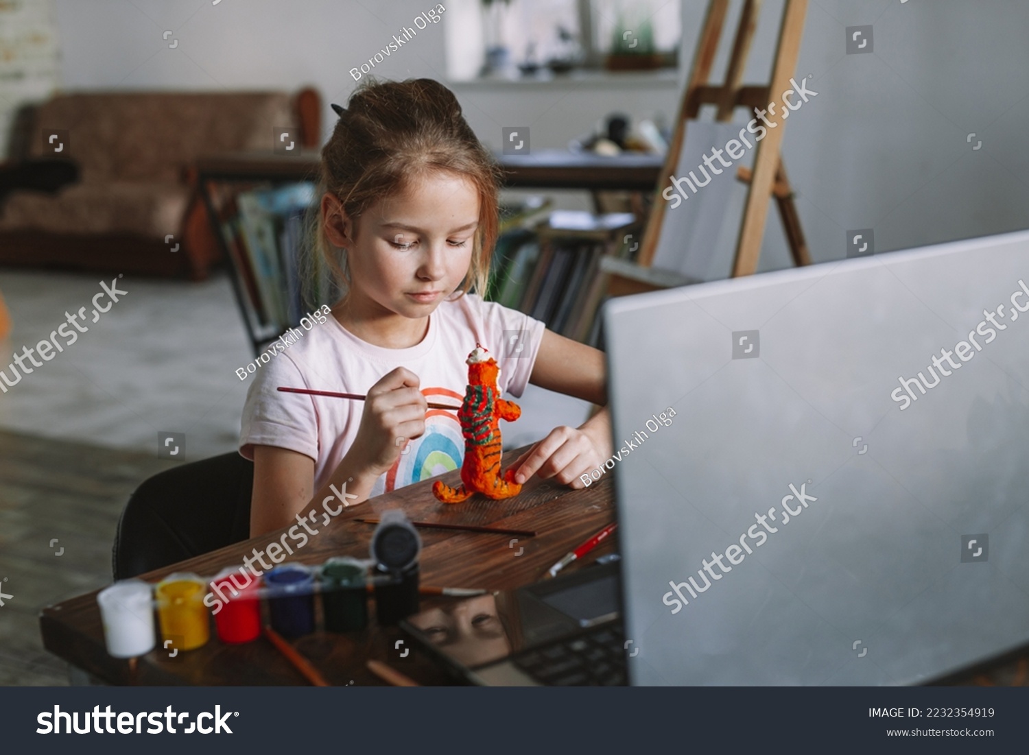 The beautiful cute girl creates crafts at home from the video lesson. A cheerful child is engaged in a hobby on a laptop. Elementary school student doing homework at homeschooling via video link. #2232354919