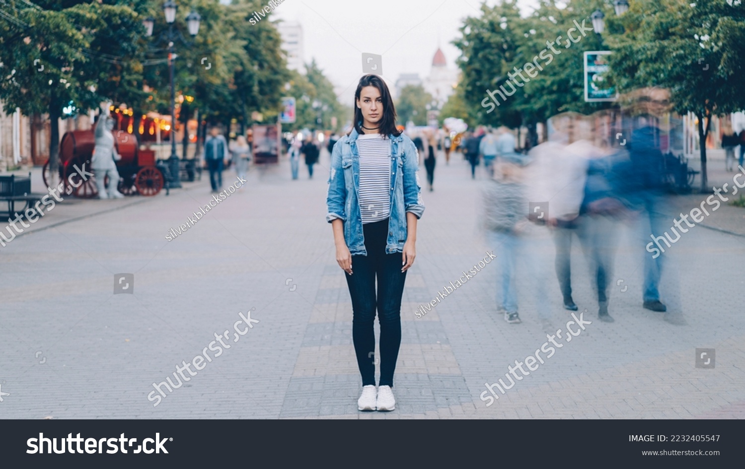 Portrait of pretty girl in trendy clothing looking at camera standing on pedestrian street by herself when busy men and women are moving around in haste. #2232405547