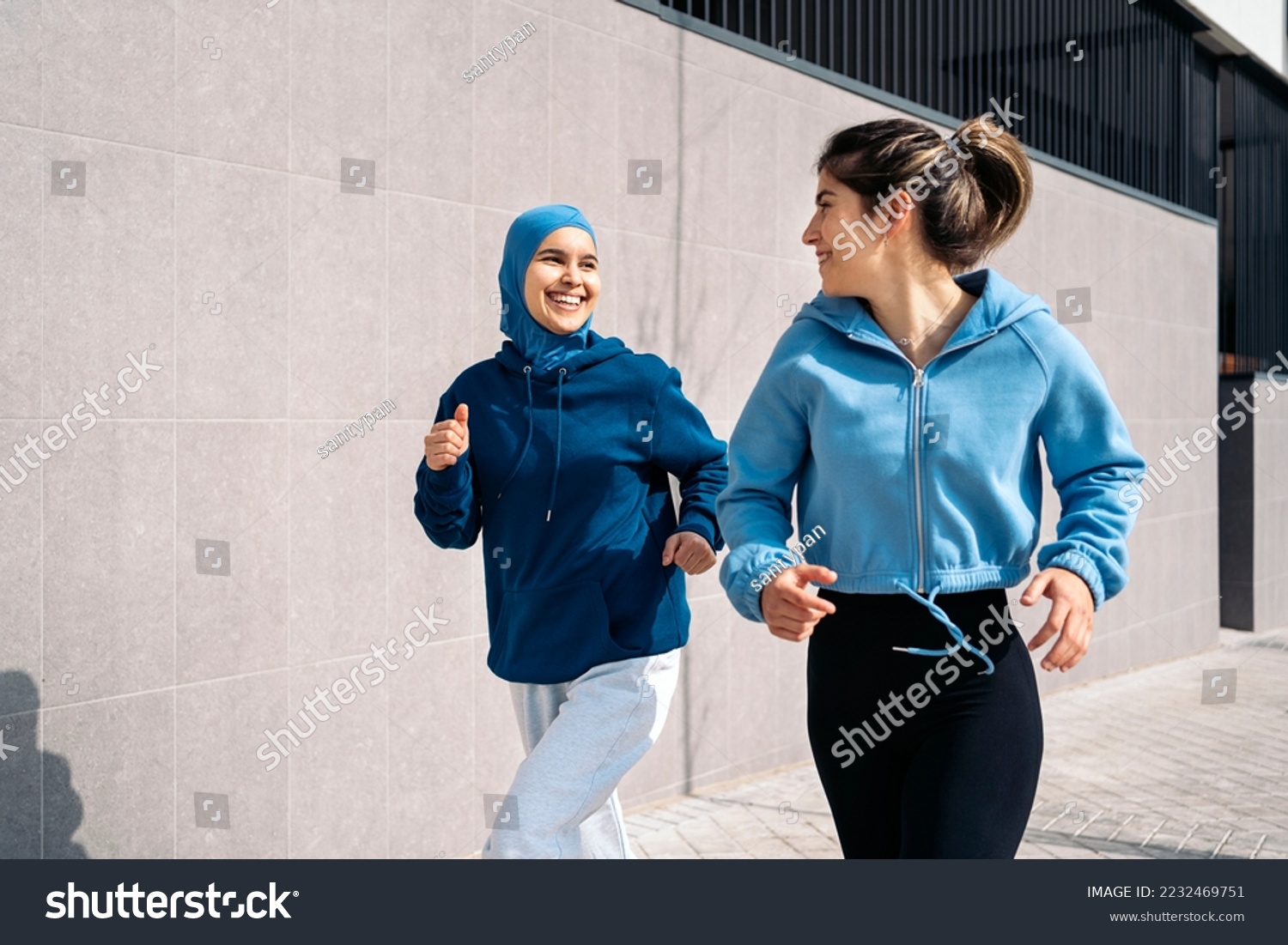 Cheerful muslim woman wearing hijab running in the street with her friend and having fun. #2232469751