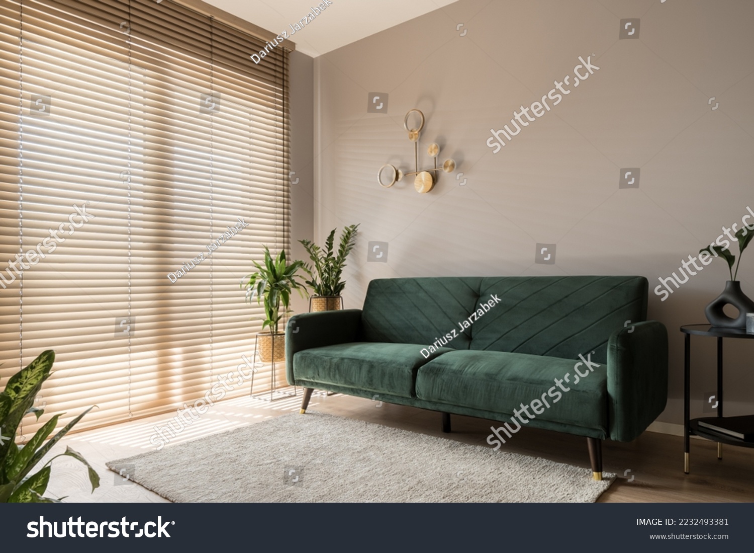 Elegant living room with big window wall behind wooden blinds, comfortable green sofa, carpet and decorative gold wall lamp #2232493381