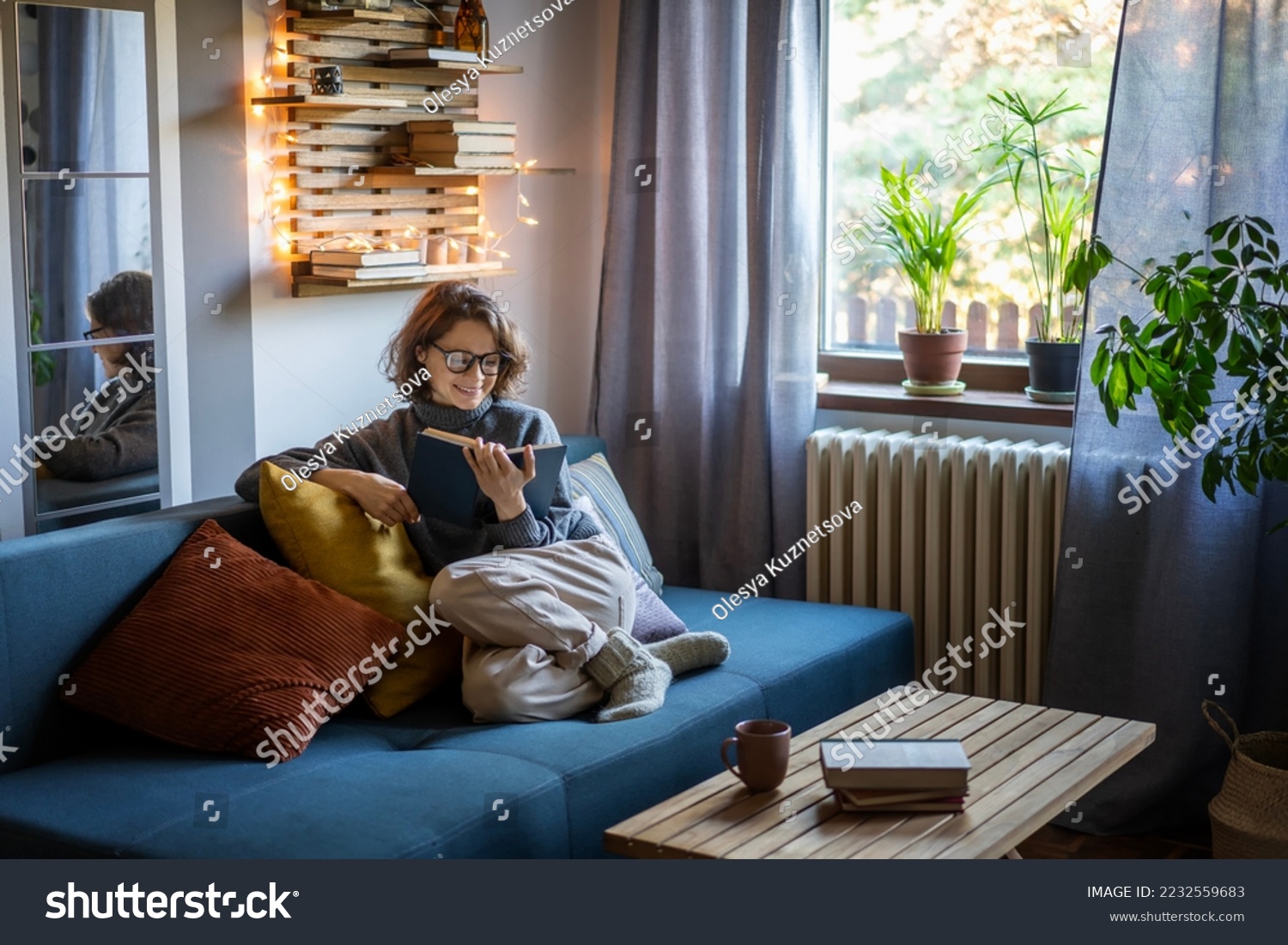 Young smiling cheerful woman in a warm sweater reading a book while sitting on the couch #2232559683