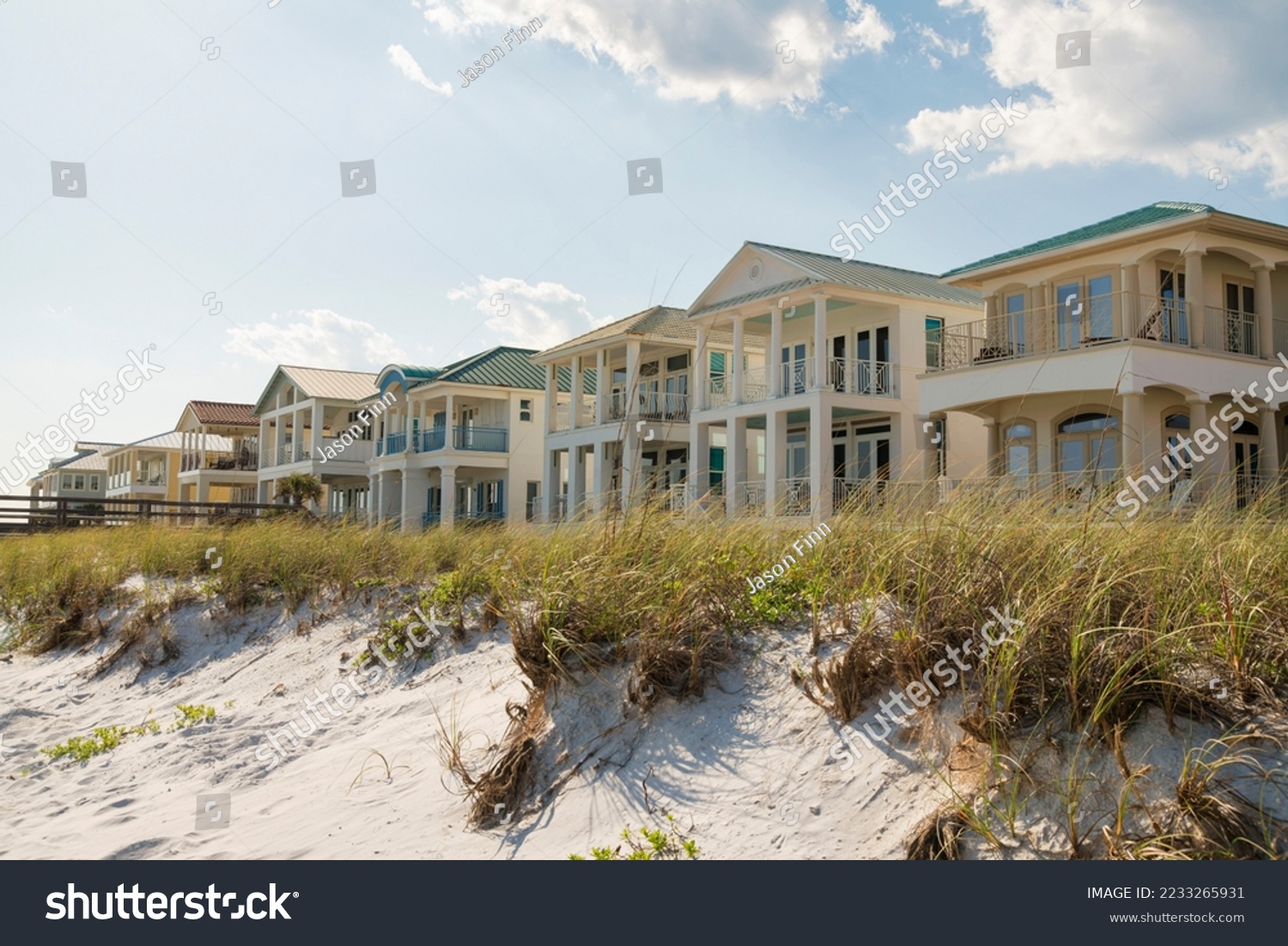 Destin, Florida- White sand dunes with grasses at the front of three-storey houses at the beach. Beach houses facade with balconies against the sky. #2233265931