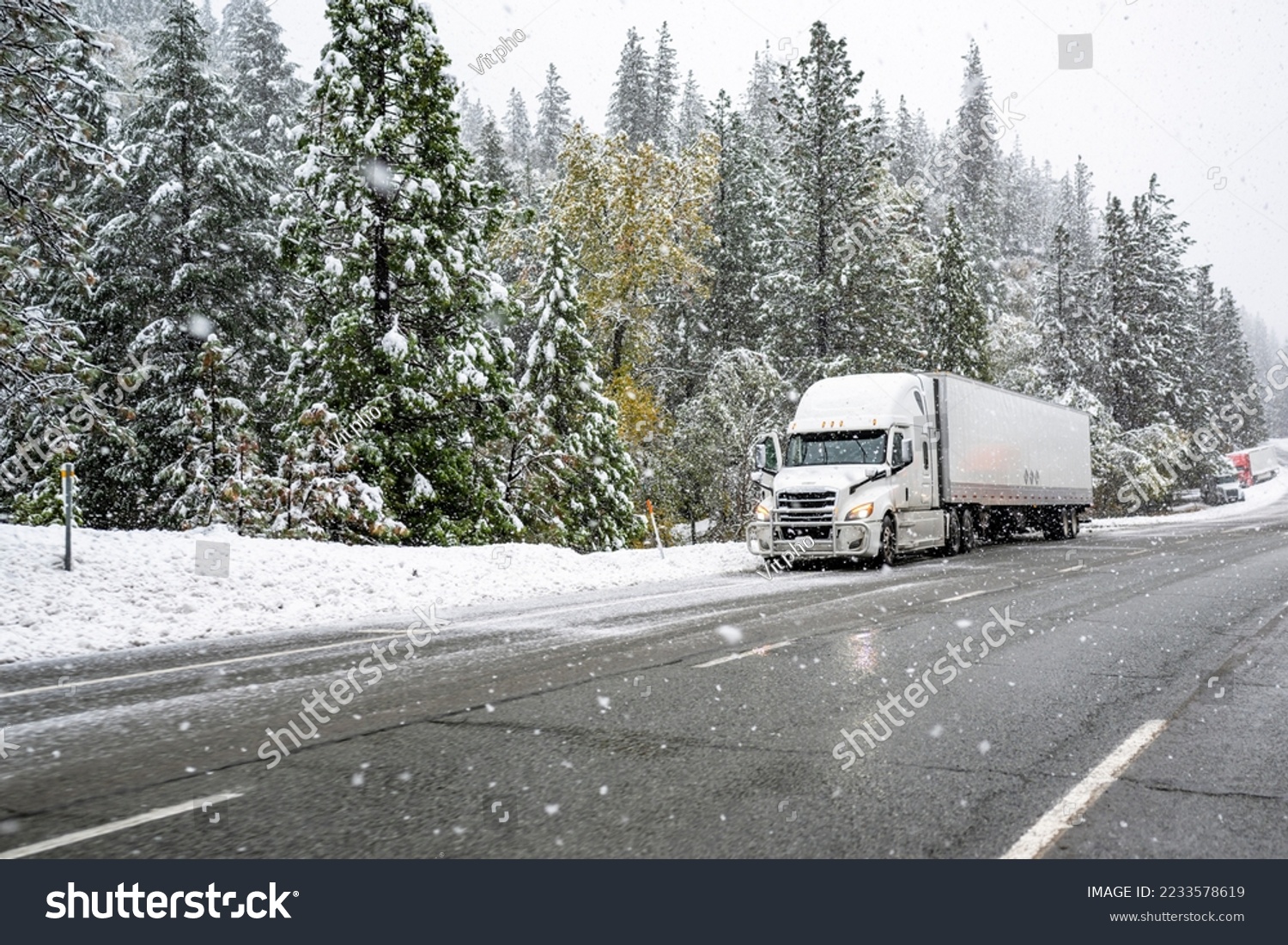 White big rig industrial semi truck with grille guard transporting cargo in dry van semi trailer standing on road shoulder of a winter highway during a snow storm near Shasta Lake in California #2233578619