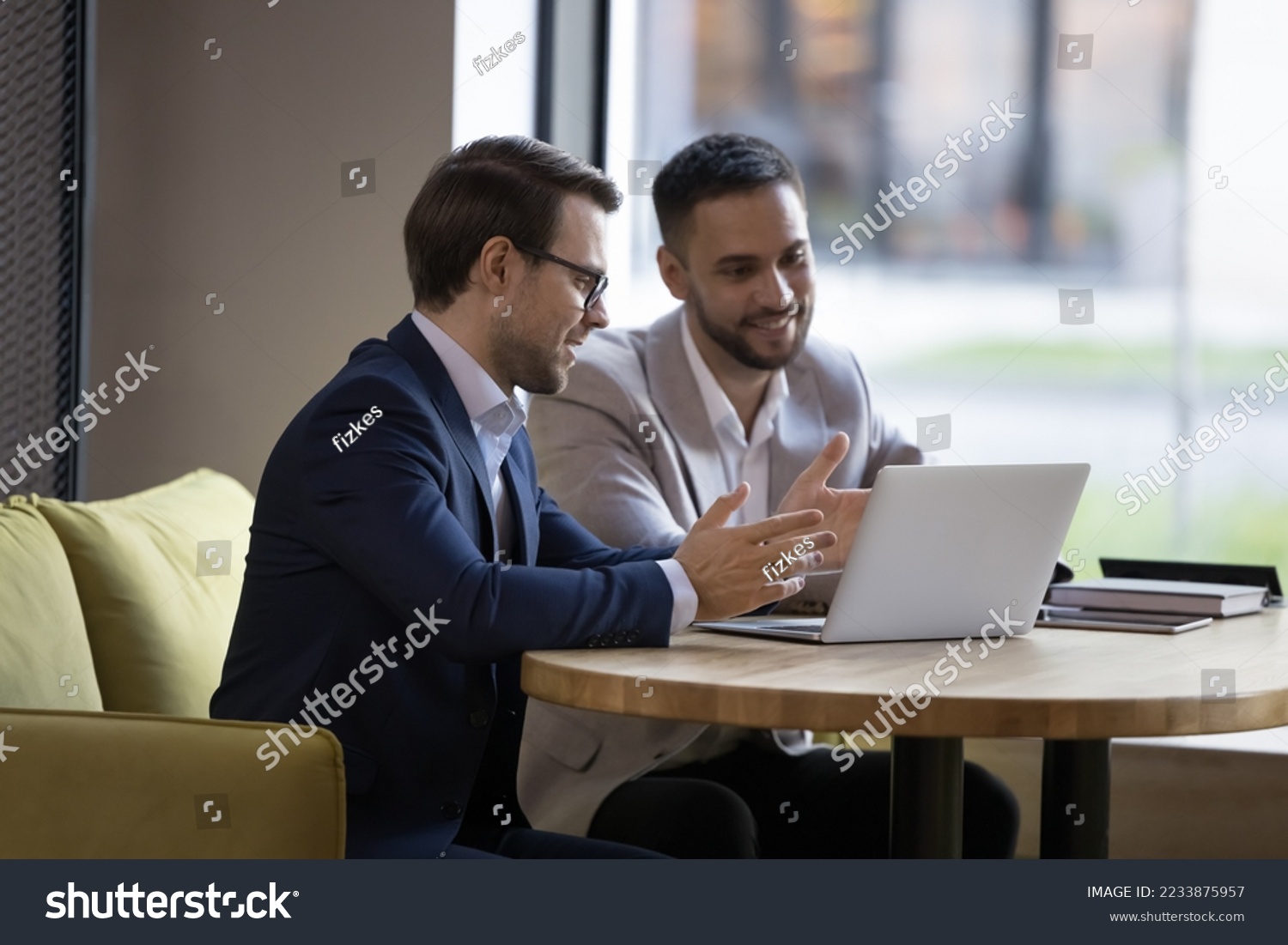 Two young businessmen in formal suits discuss new software sit at table with laptop in workspace, work on startup project, manager offering, presenting new services to client on device meet in office #2233875957