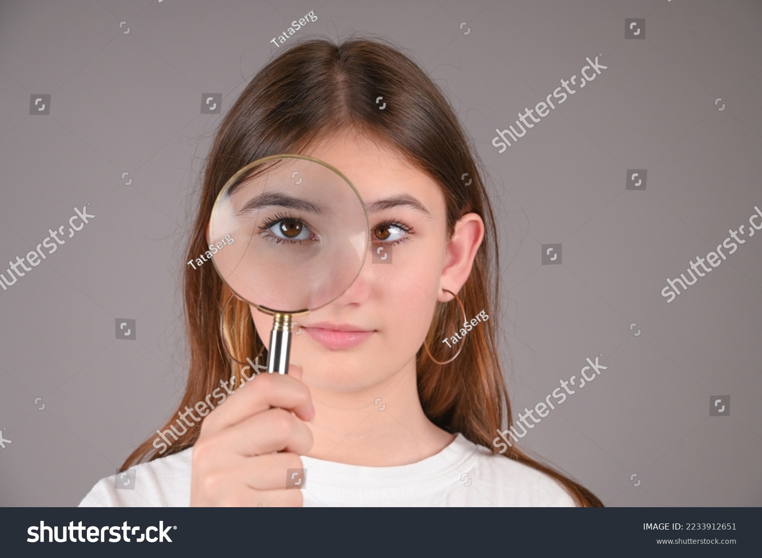 Teenage girl with magnifying glass on gray background. Female eyes with strabismus.  #2233912651