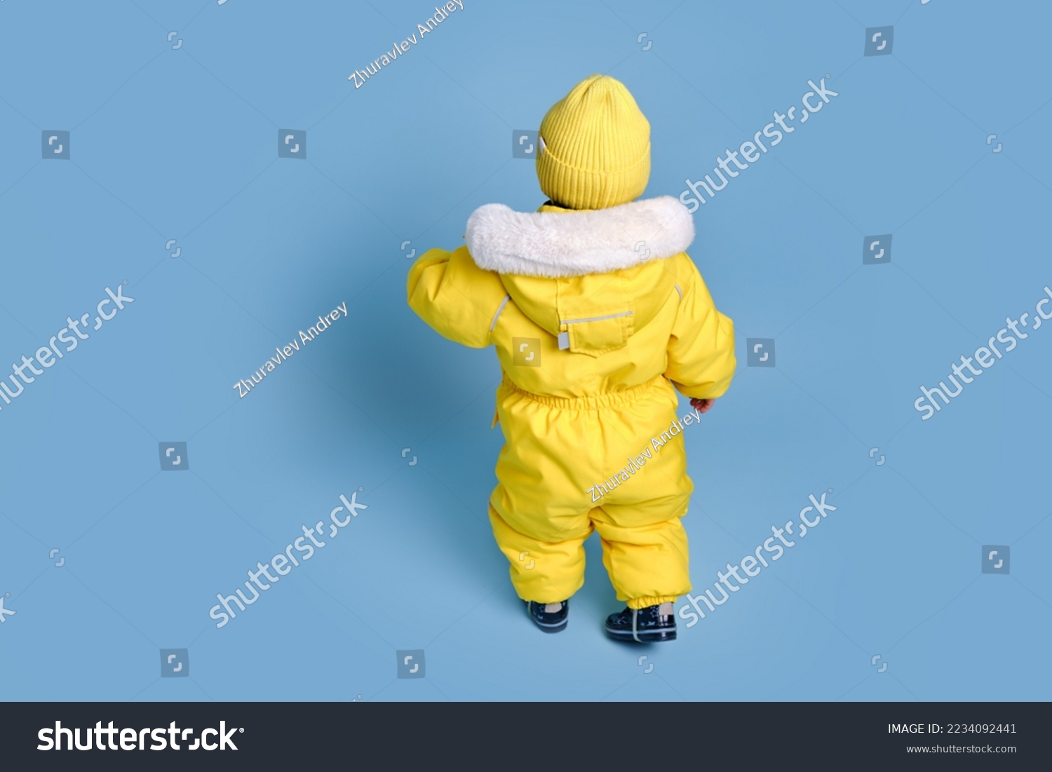 Happy toddler baby in winter clothes snowsuit on studio blue background. A child in a warm yellow jumpsuit with a hood. Kid aged one year five months #2234092441