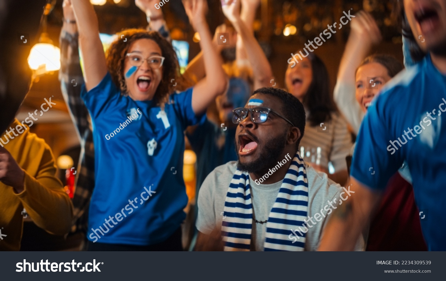 Group of Multiethnic Friends Watching a Live Soccer Match on TV in a Sports Bar. Fans with Painted Faces Cheering. Young People Celebrating When Team Scores a Goal and Wins the Football World Cup. #2234309539