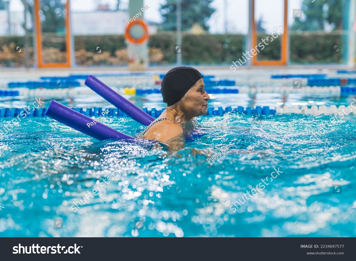 Side view indoor portrait focused caucasian senior woman in a black head cap using violet pool noodle during swimming. Equipment concept. High quality photo #2234697577