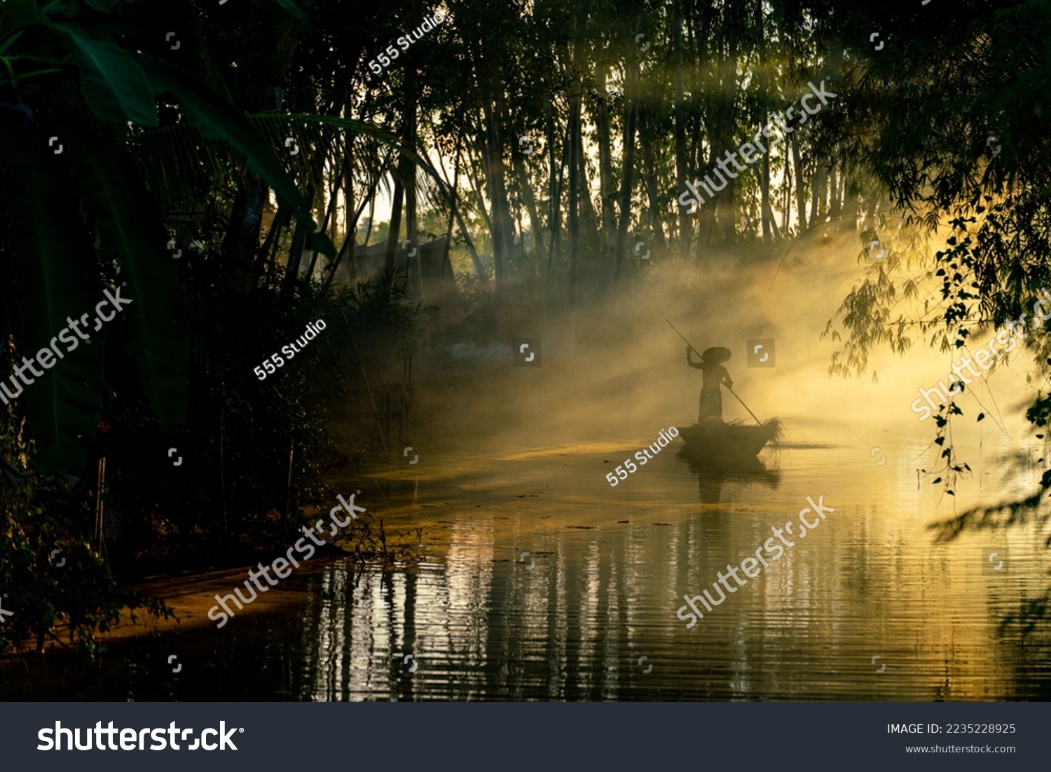 Beautiful view of an asian adult male old boatman rowing a wooden boat with a bamboo stick across a small river stream during sunset to deliver dry grasses for animal feeds in northeast Thailand #2235228925