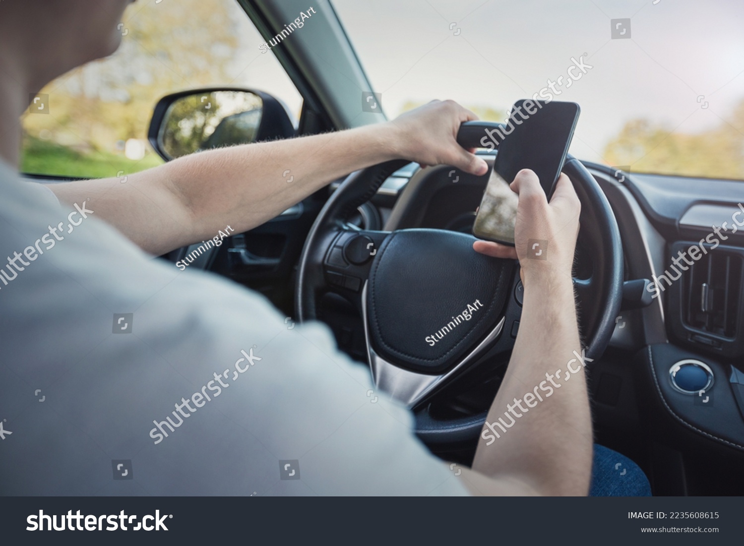 Young driver guy distracted by his phone while in front of the steering wheel, using his smartphone with one hand while driving. Risk and danger situations on the road, violating traffic rules #2235608615
