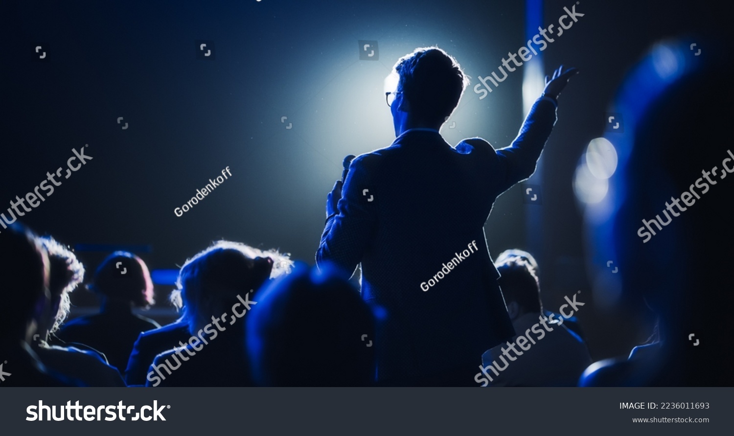 Backview of a Stylish Young Businessman in a Dark Crowded Auditorium at a Startup Summit. Young Man Talking to a Microphone During a Q and A session. Entrepreneur Happy with Event Speaker. #2236011693