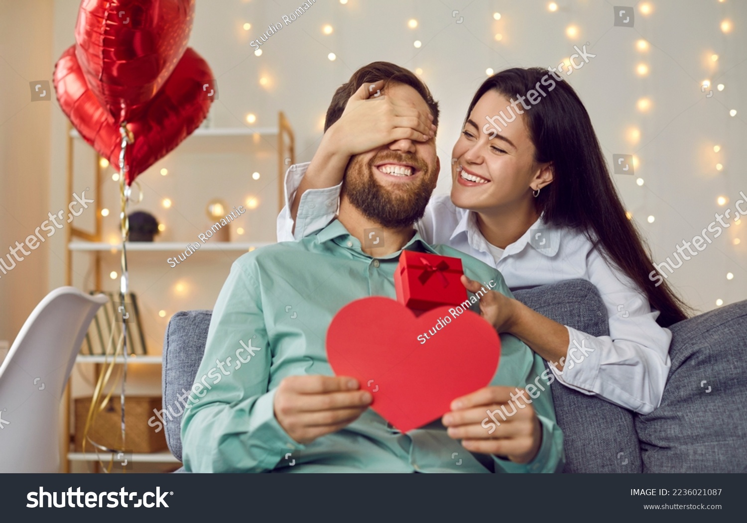 Joyful young couple celebrating St Valentine's Day. Loving woman makes surprise for her boyfriend on Saint Valentine's Day. Happy girlfriend covers man's eyes and gives him greeting card and gift box #2236021087