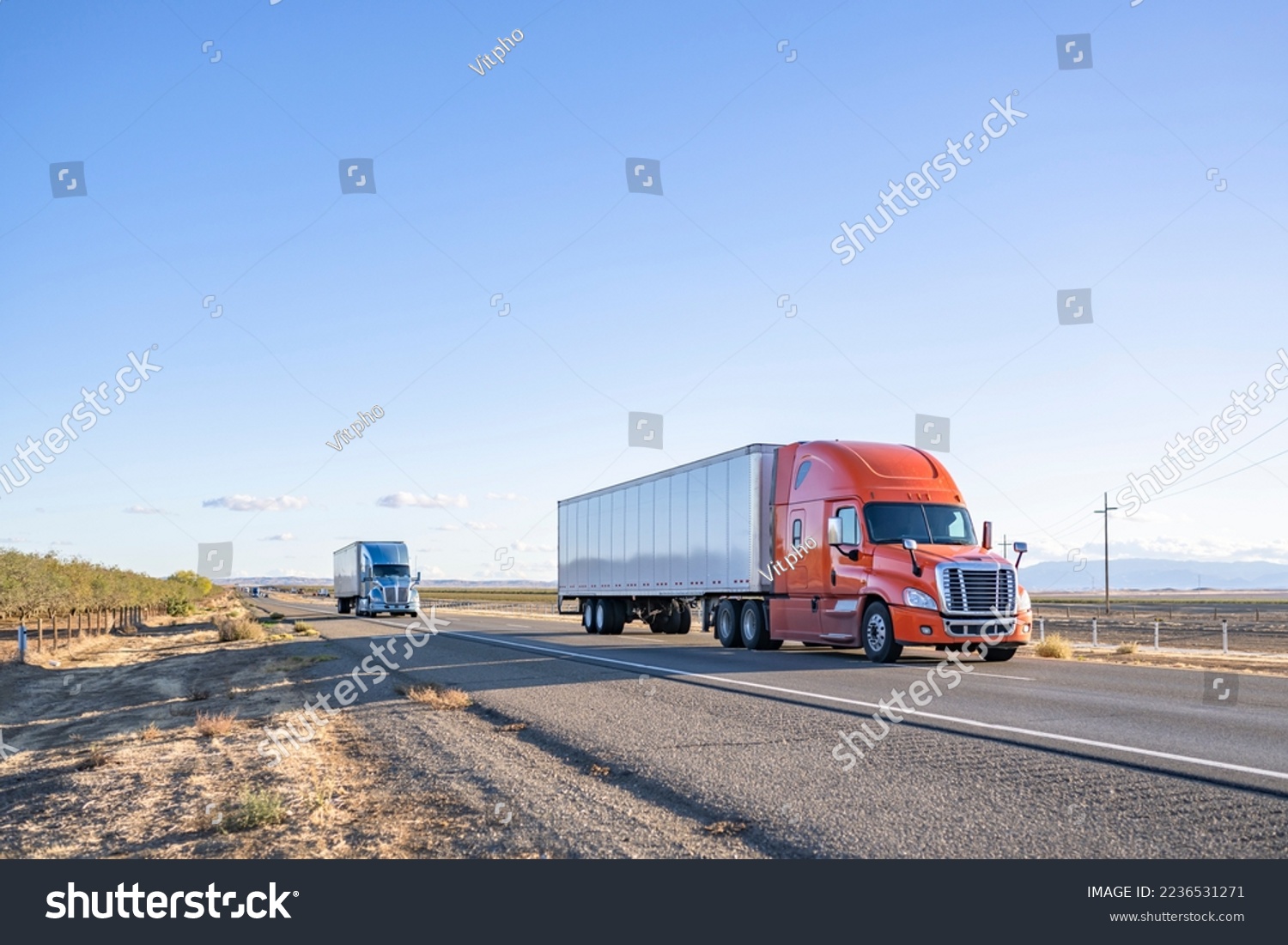 Industrial long hauler big rigs semi trucks team transporting commercial cargo in loaded dry van semi trailers running together on the flat straight highway road in California #2236531271