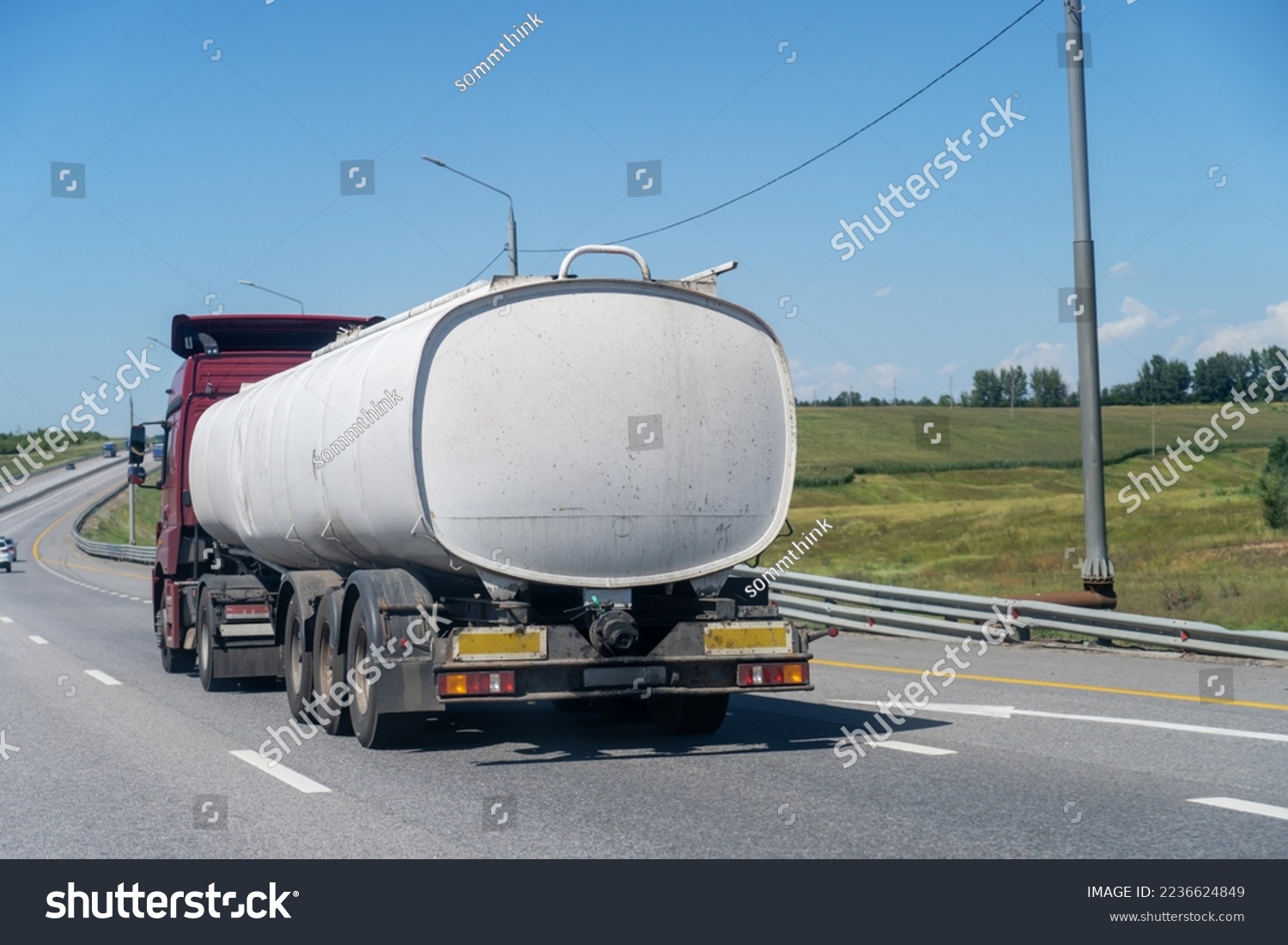 Red truck carrying tank semi-trailer on country road. Modern truck pulling white tank trailerswith combustible material through countryside in summer #2236624849