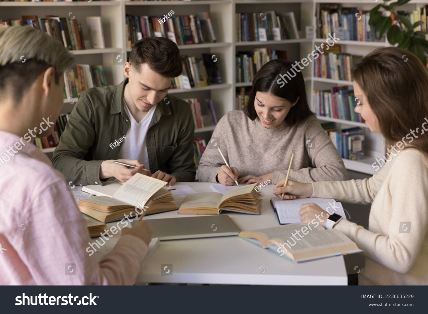 Group of positive young hardworking smart students studying in library, sitting together at open books, writing notes, essay, talking, working on team task, research, preparing report #2236635229