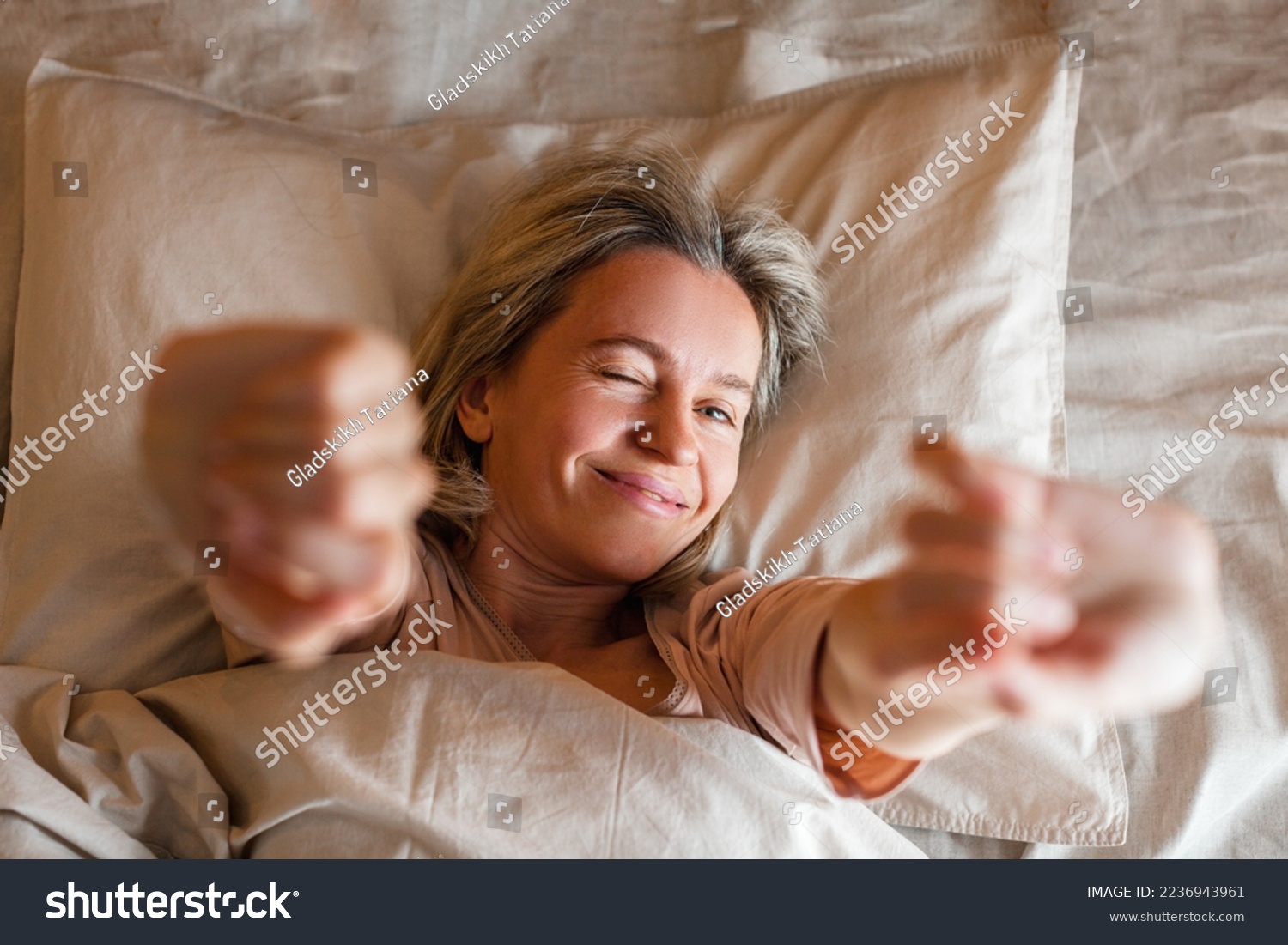 Good morning, new day, weekend, holiday. Happy middle aged woman sits on bed, lady stretching arms after sleep and enjoying morning in cozy comfort bedroom interior, free space #2236943961