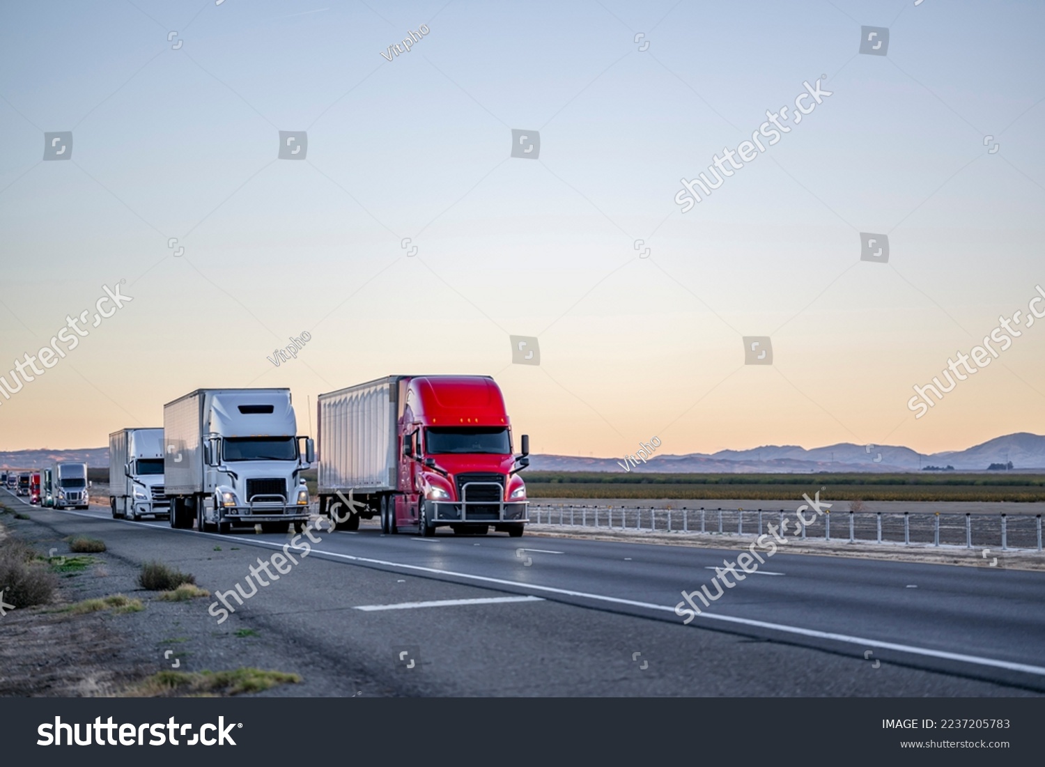Convoy of a industrial grade commercial professional transportation of different big rig semi trucks with semi trailers driving on the straight highway road at twilight time in California #2237205783