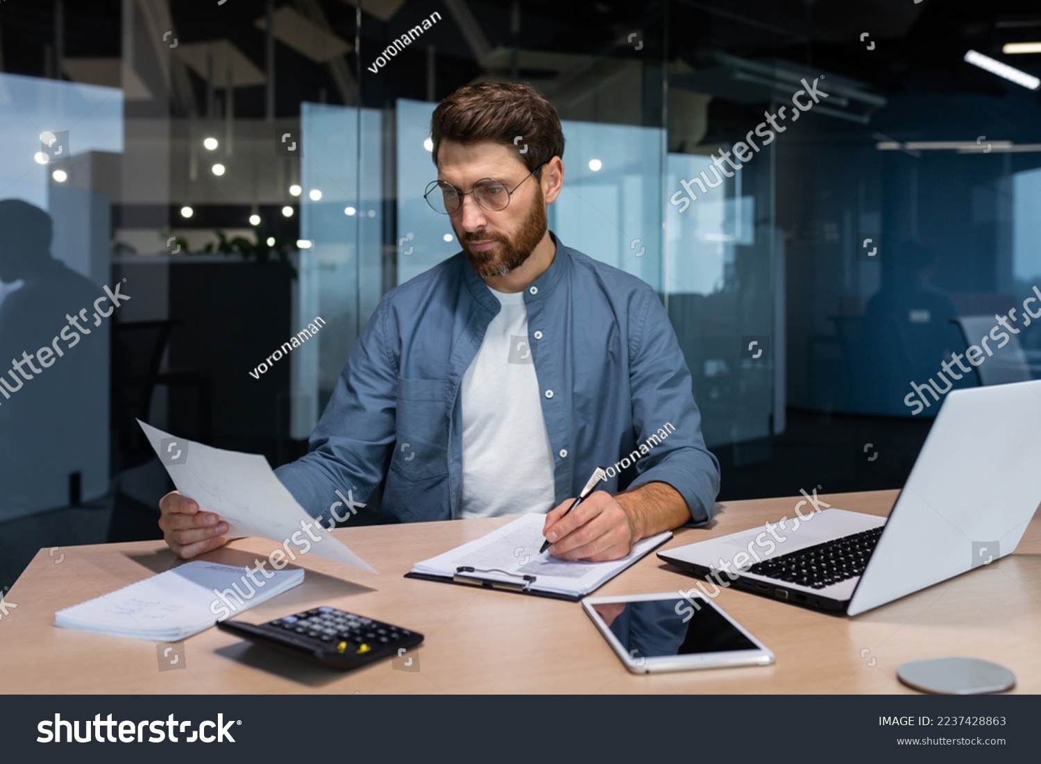 Serious and focused financier accountant on paper work inside office, mature man using calculator and laptop for calculating reports and summarizing accounts, businessman at work in casual clothes. #2237428863