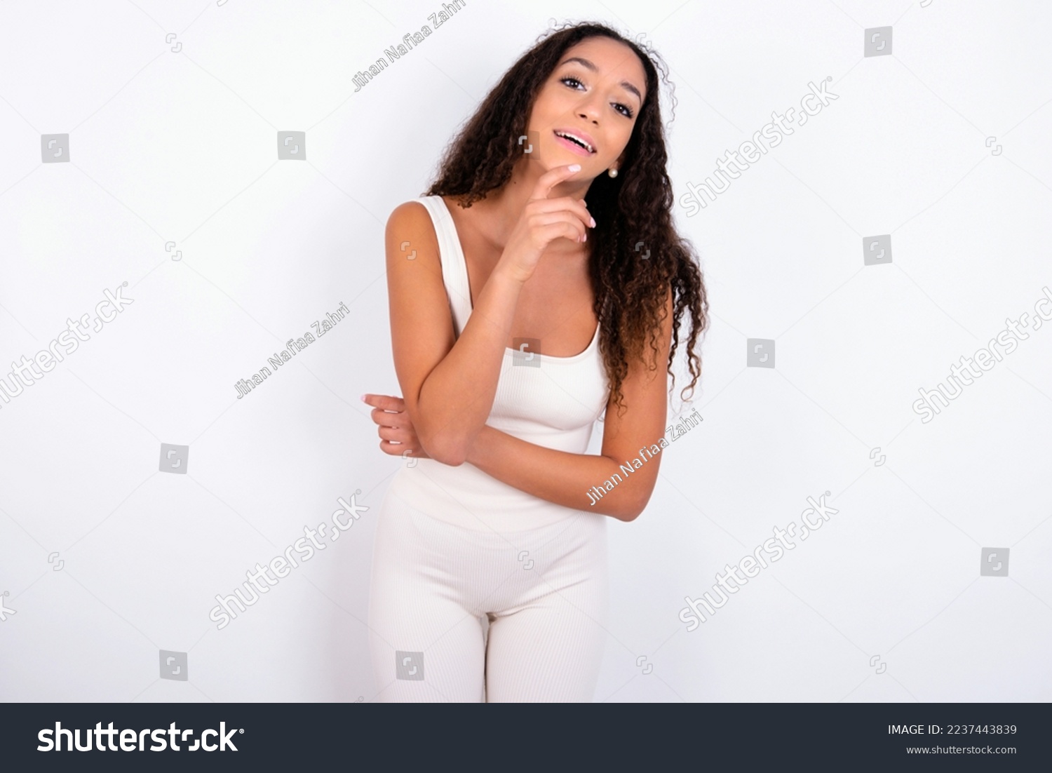 teen girl with curly hair wearing white sport set over white background laughs happily keeps hand on chin expresses positive emotions smiles broadly has carefree expression #2237443839
