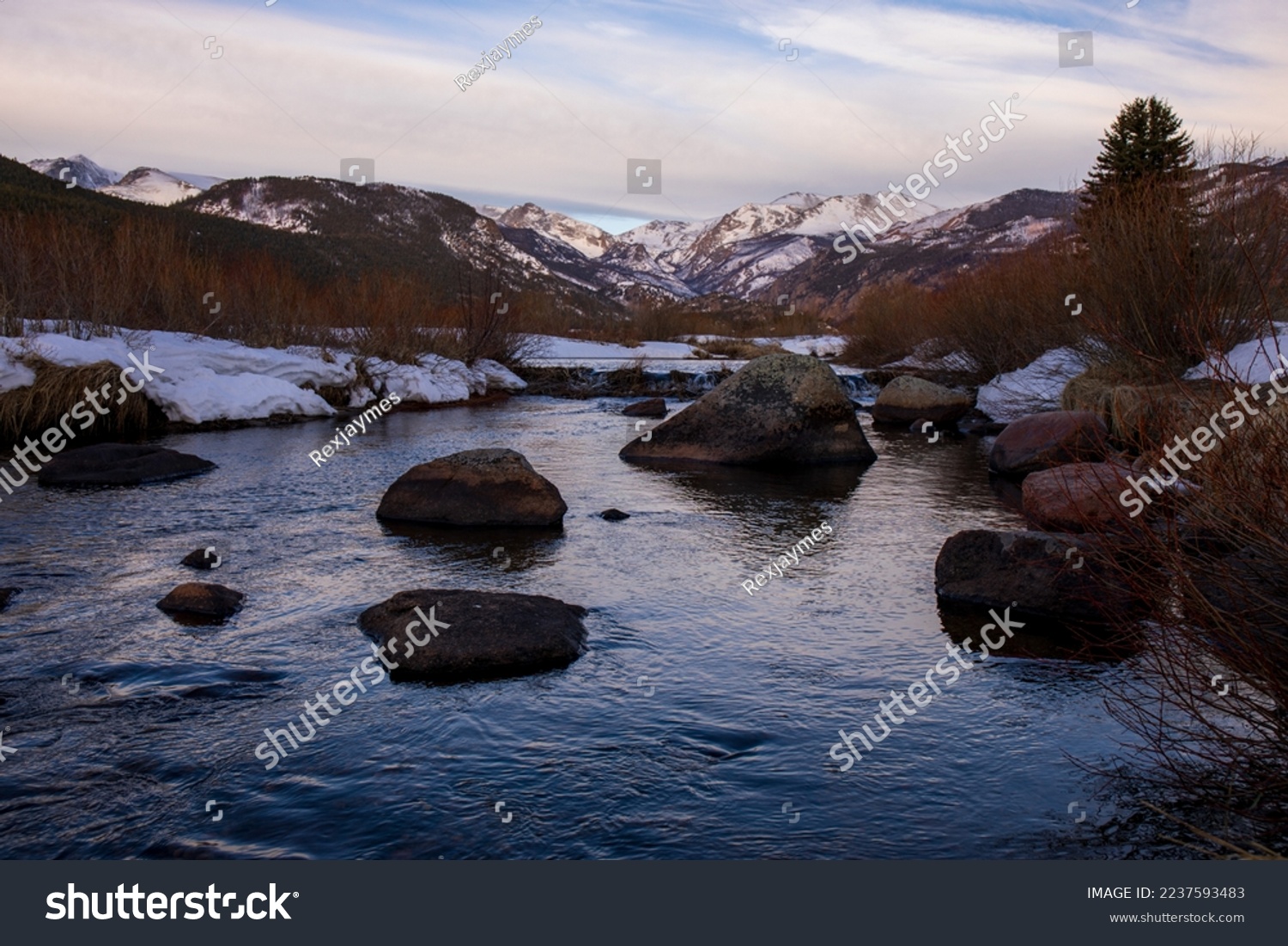 Snow covered river banks in nature setting with a slow flowing stream and a pale sky.  Rocky Mountain stream with blue water, dormant grass.  Colorado high country with cloud filled sunrise sky. #2237593483