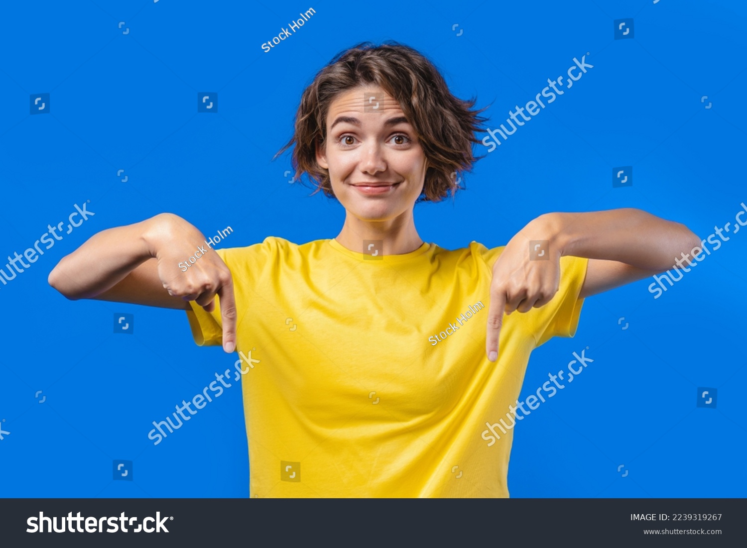 Pretty woman pointing down to advertising area. Blue background. Young lady asking to click to subscribe below. Copy space for your commercial idea, promotional content. High quality photo #2239319267