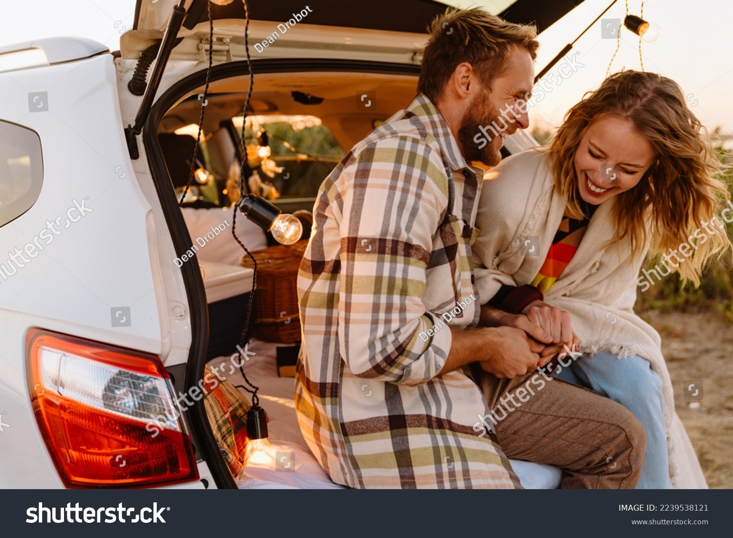 Happy young white couple smiling and sitting in car trunk together outdoors #2239538121