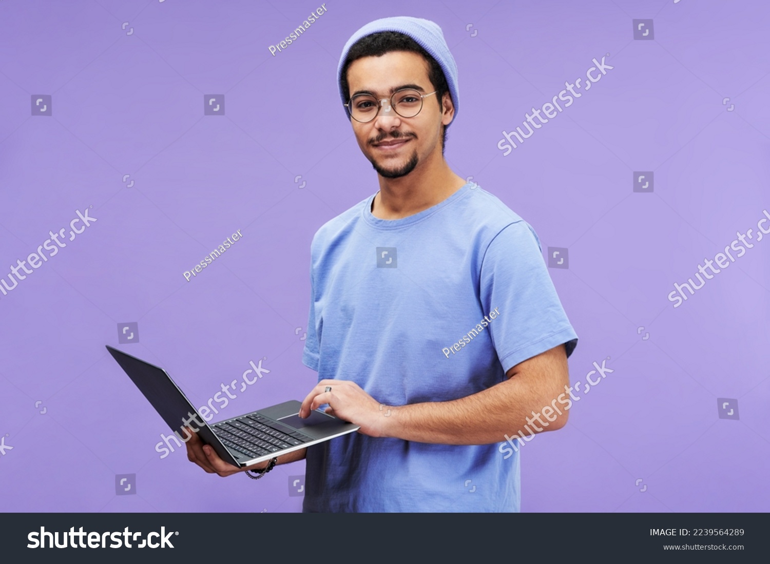 Young successful student or designer in blue t-shirt and beanie hat holding laptop while looking at camera against lavender background #2239564289