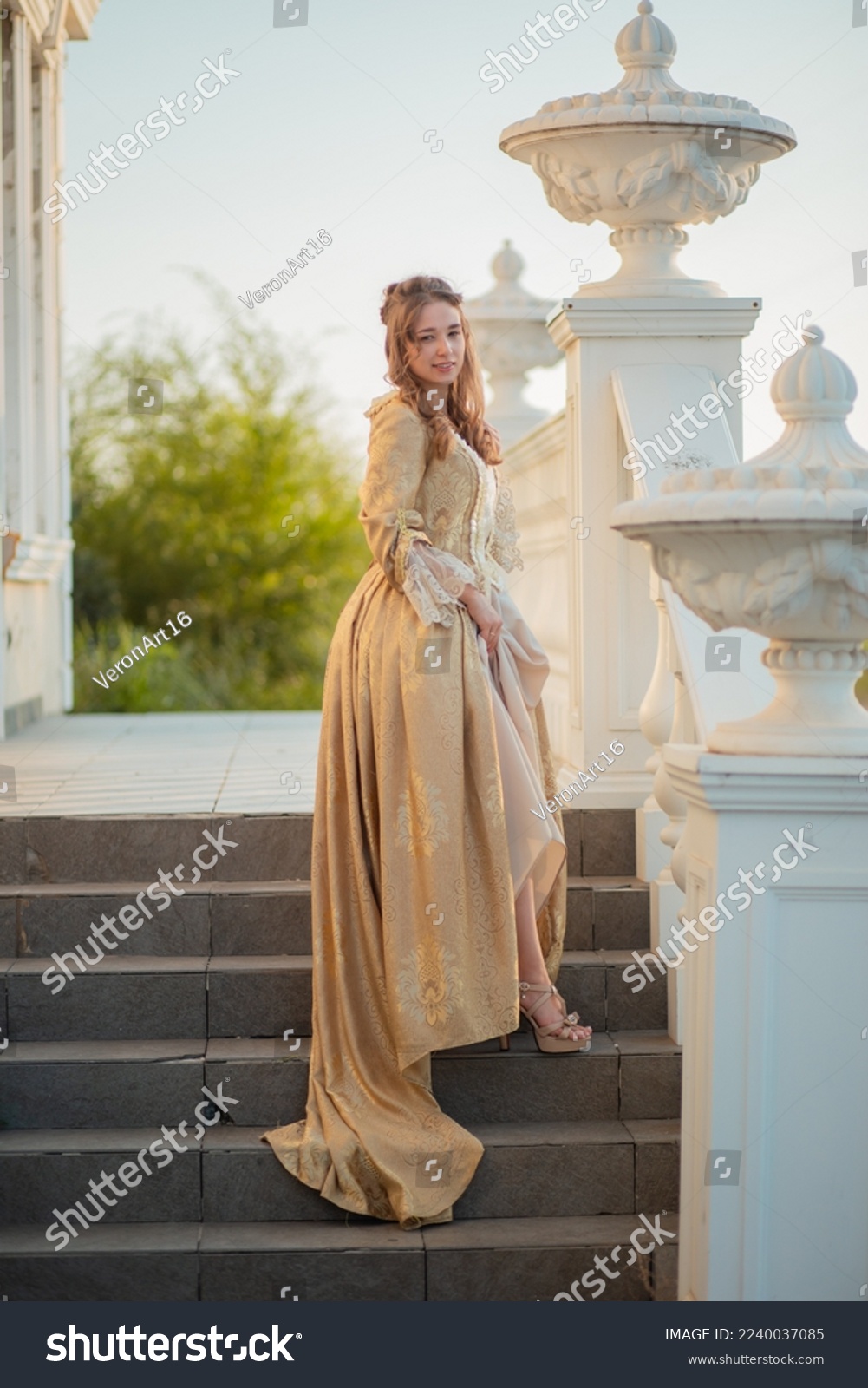 A beautiful young woman in a historic eighteenth century gold dress stands on the stairs of the mansion. Princess in the palace. fabulous image #2240037085