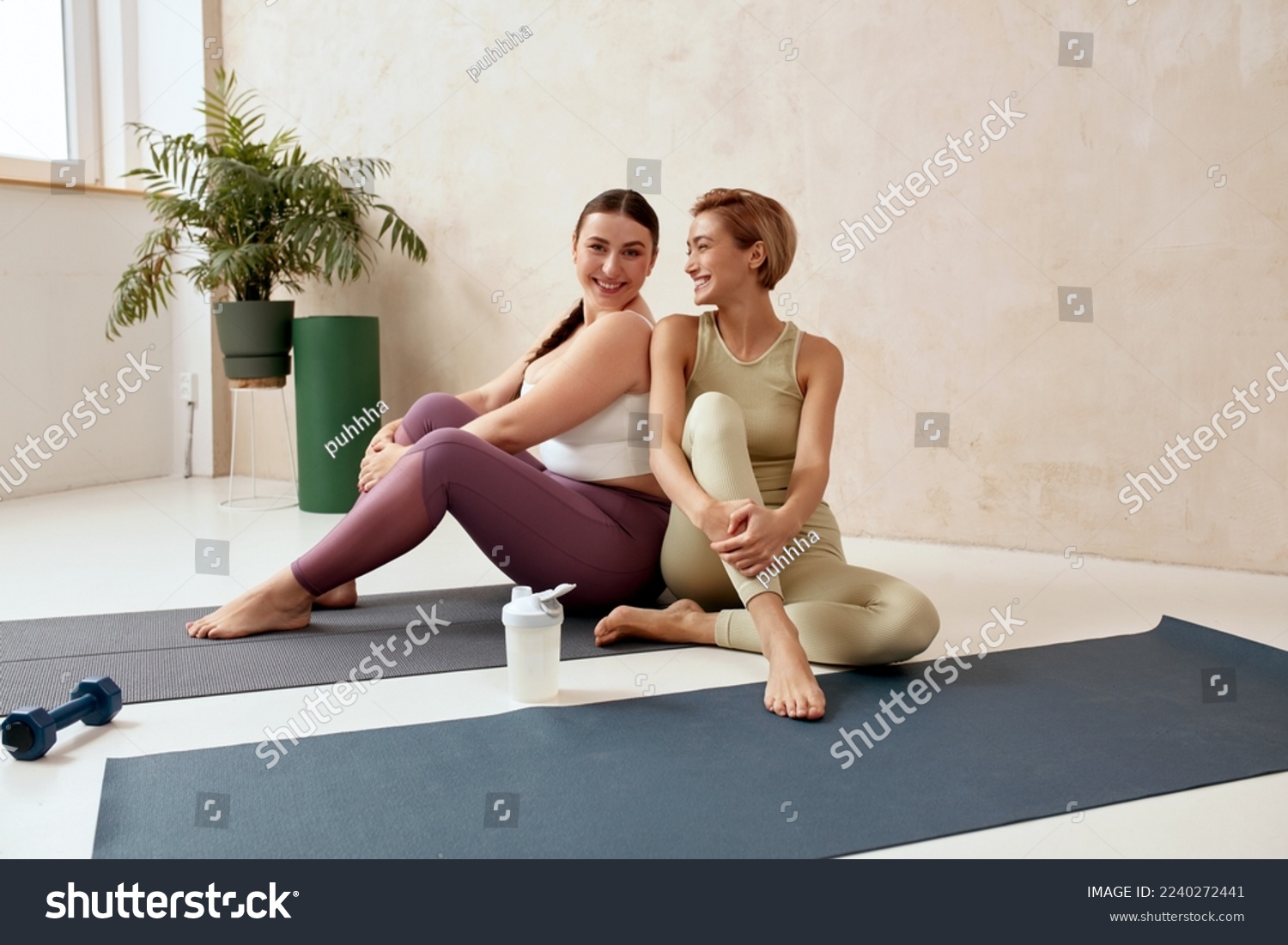 Smiling Woman Embracing After Yoga. Female Friends Posing After Training Together At Home. Attractive Trainer And Her Client In Sportswear Sitting On Mat #2240272441