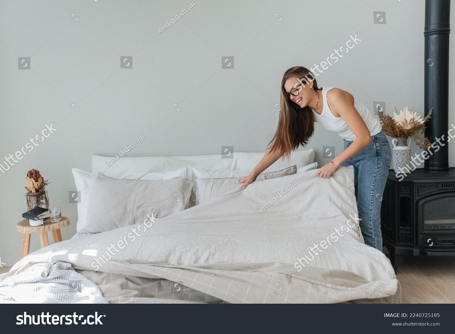 Laughing caucasian beautiful young woman makes bed wears jeans and white t-shirt and glasses. Pretty hispanic girl makes housekeeping. Mockup, people at home. Cheerful student makes order home. #2240725195