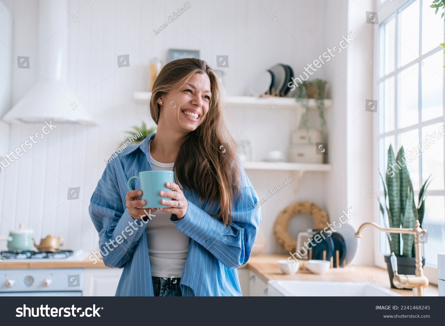 Happy caucasian female with long loose hair in jeans and blue shirt toothy smiles holds cup of tea looks aside happily enjoys weekend at cozy home. Pretty hispanic woman laughs against blurry kitchen. #2241468245
