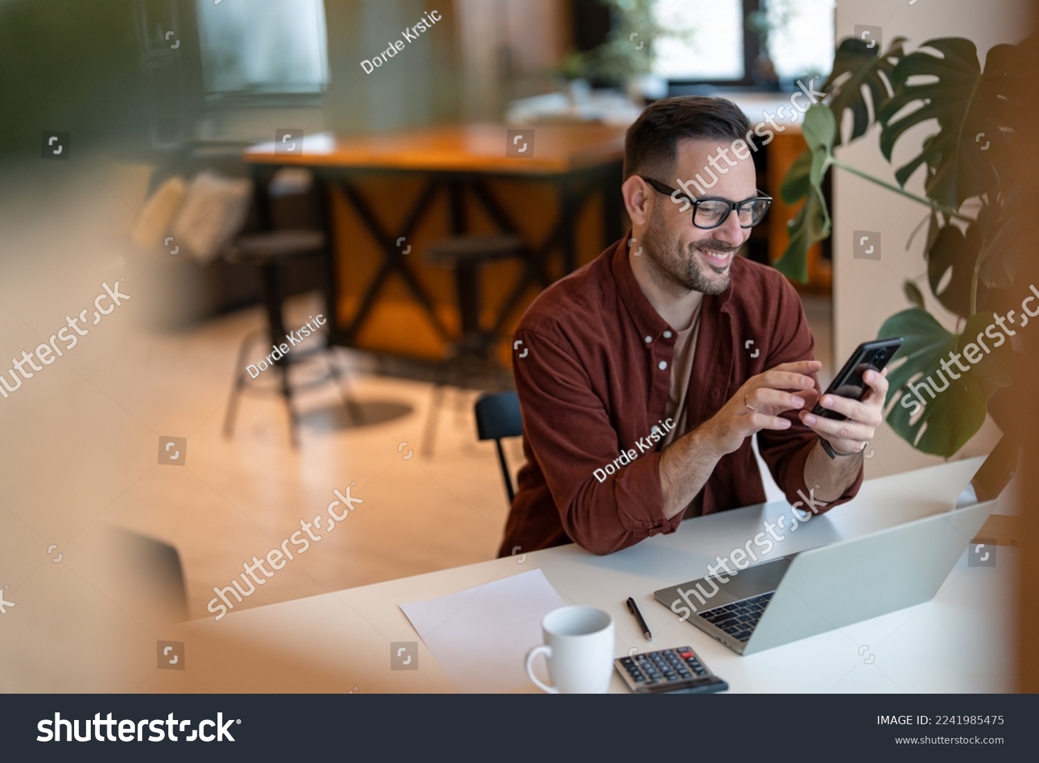 Shot of a young businessman using a smart phone in a home office. Smiling, touching the screen, browsing the internet. #2241985475
