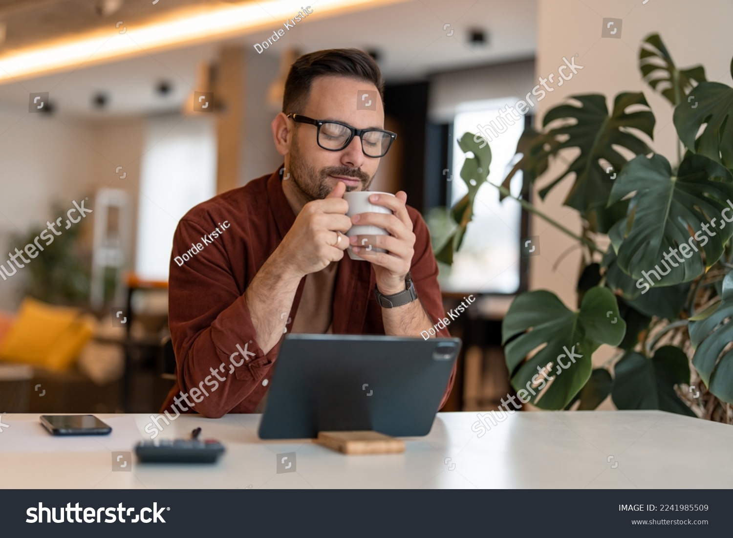 Photo of an adult man drinking coffee while having a break from work. Young content freelancer having a coffee, daydreaming with his eyes closed, smelling coffee, enjoying his break in a home office. #2241985509