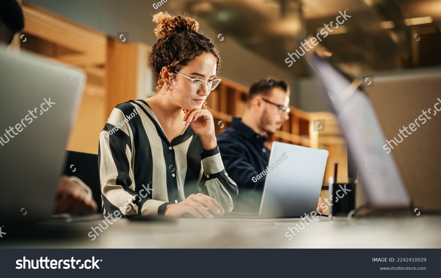 Portrait of Enthusiastic Hispanic Young Woman Working on Computer in a Modern Bright Office. Confident Human Resources Agent Smiling Happily While Collaborating Online with Colleagues. #2242410029