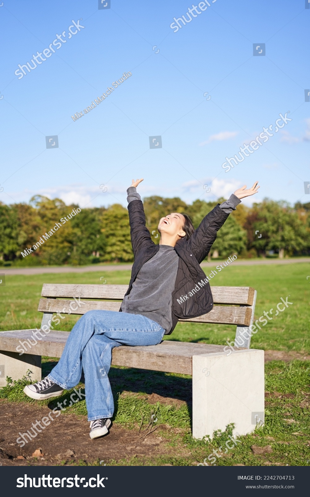 Happy asian woman stretching her hands, sitting on bench with excited face, smiling pleased, feeling freedom, enjoying day in park. #2242704713