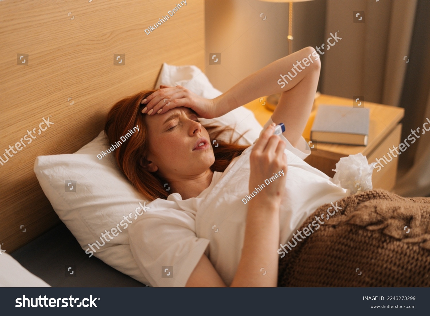 Illness tired woman touching forehead after using thermometer to checking body temperature and take medicine lying under blanket on bed. Sick female with cold lying in bed with high fever in bedroom. #2243273299