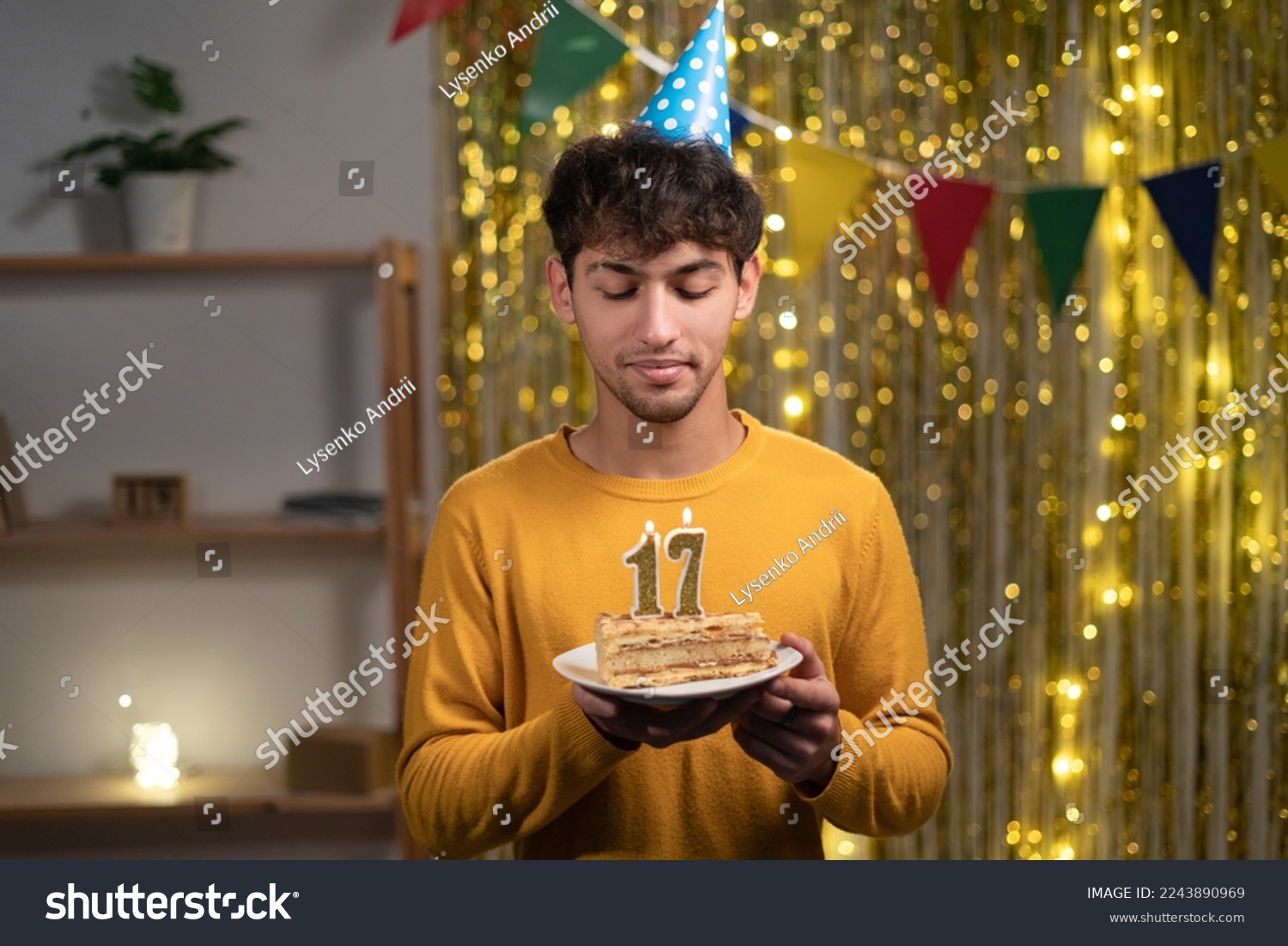 Young man in cone party hat to make wish holding his birthday cake with candles number 17 during celebration at home. Seventeens birthday concept #2243890969