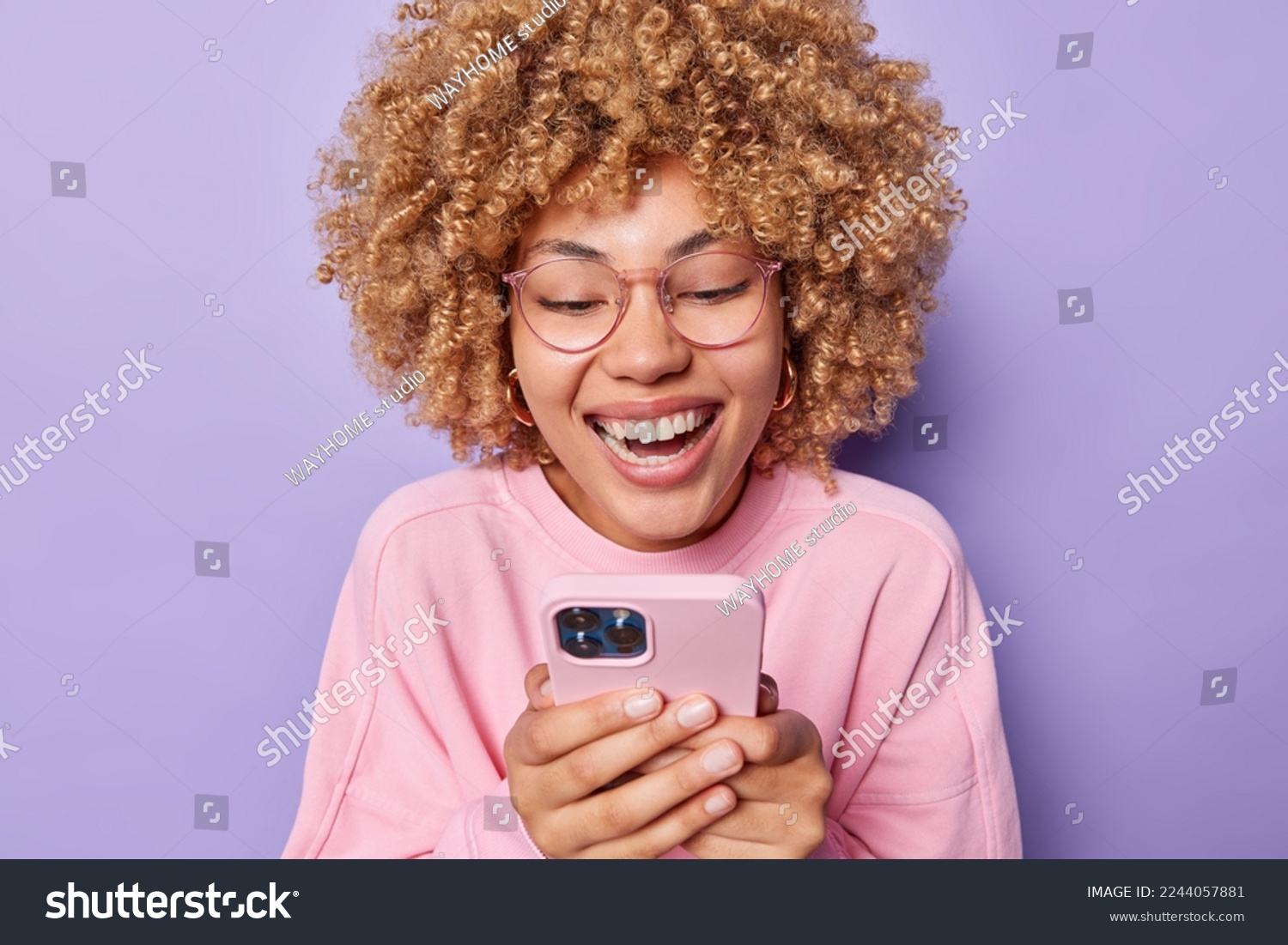 Positive curly haired young woman smiles happily and focused at smartphone screen reads funny story dressed in casual pink pullover wears transparent eyeglasses isolated over purple background. #2244057881