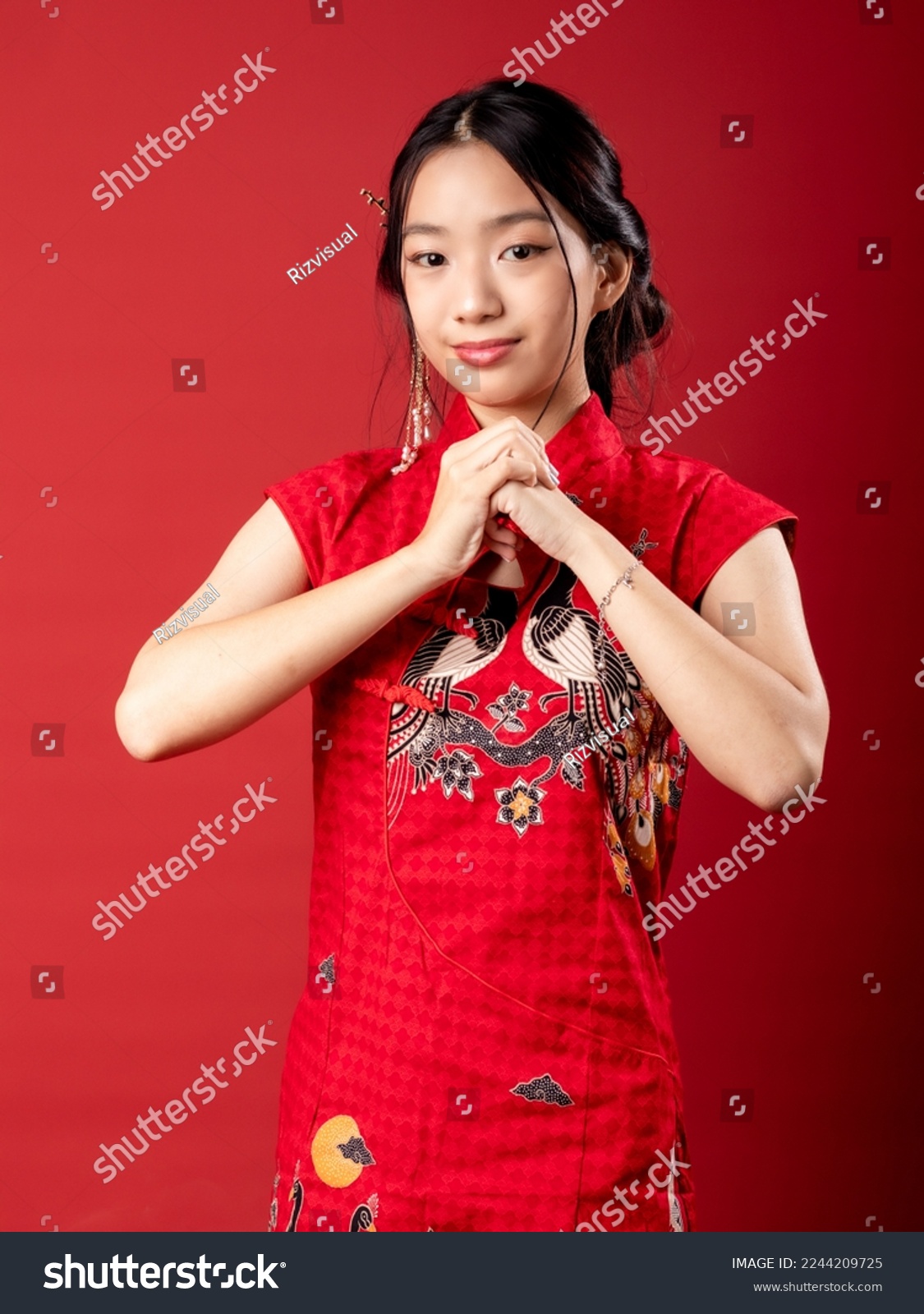 A 20-year-old Chinese Indonesian (Asian) girl in a Chinese traditional red dress (Cheongsam), with her hand, folded over each other as a gesture to greet Happy CNY. Isolated on a red background. #2244209725