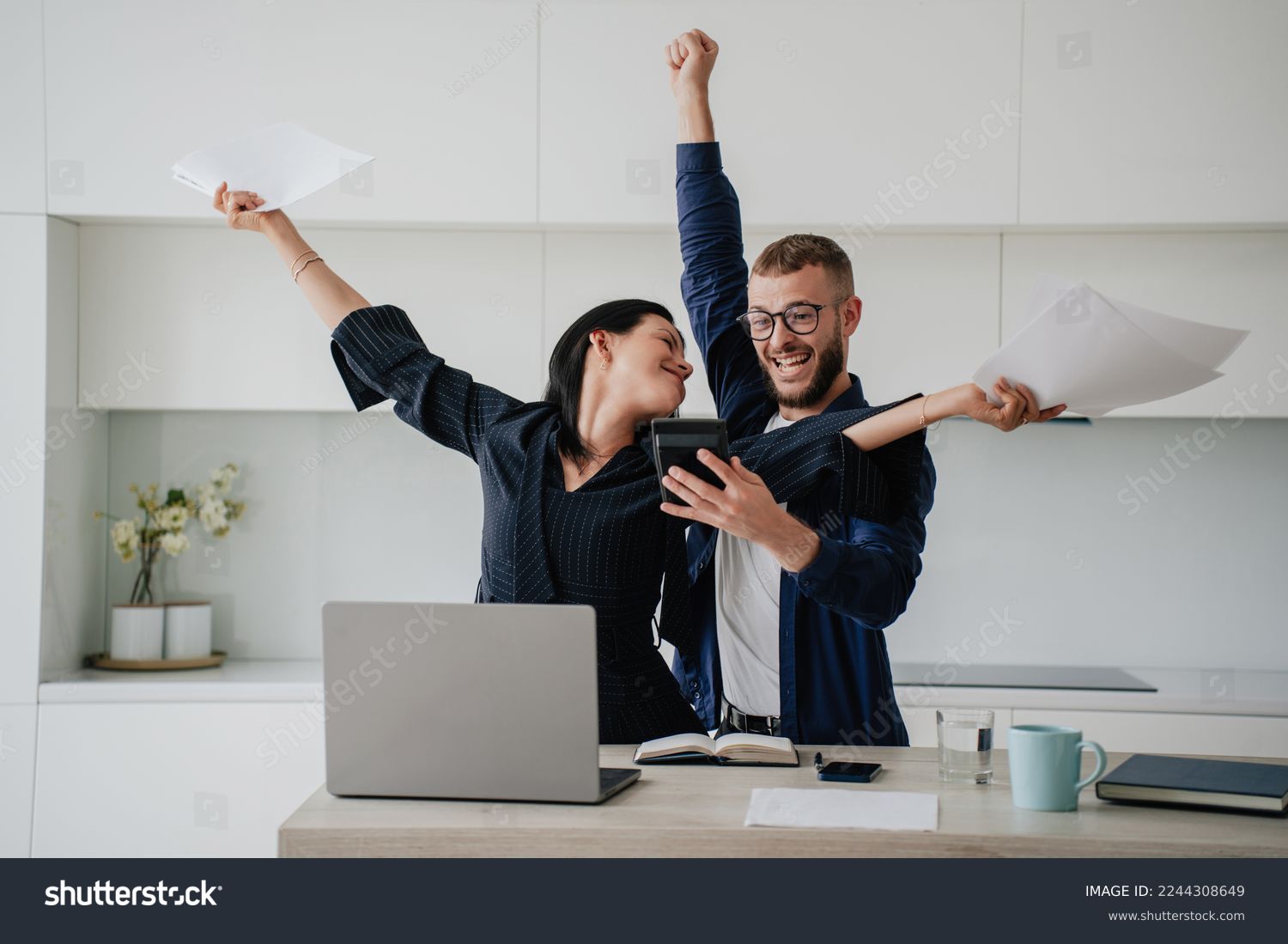 Excited caucasian couple after calculate their year earnings, celebrating successful business at new home using calculator, laptop. Woman raises hands in winner gesture holds papers. Financial freedom #2244308649