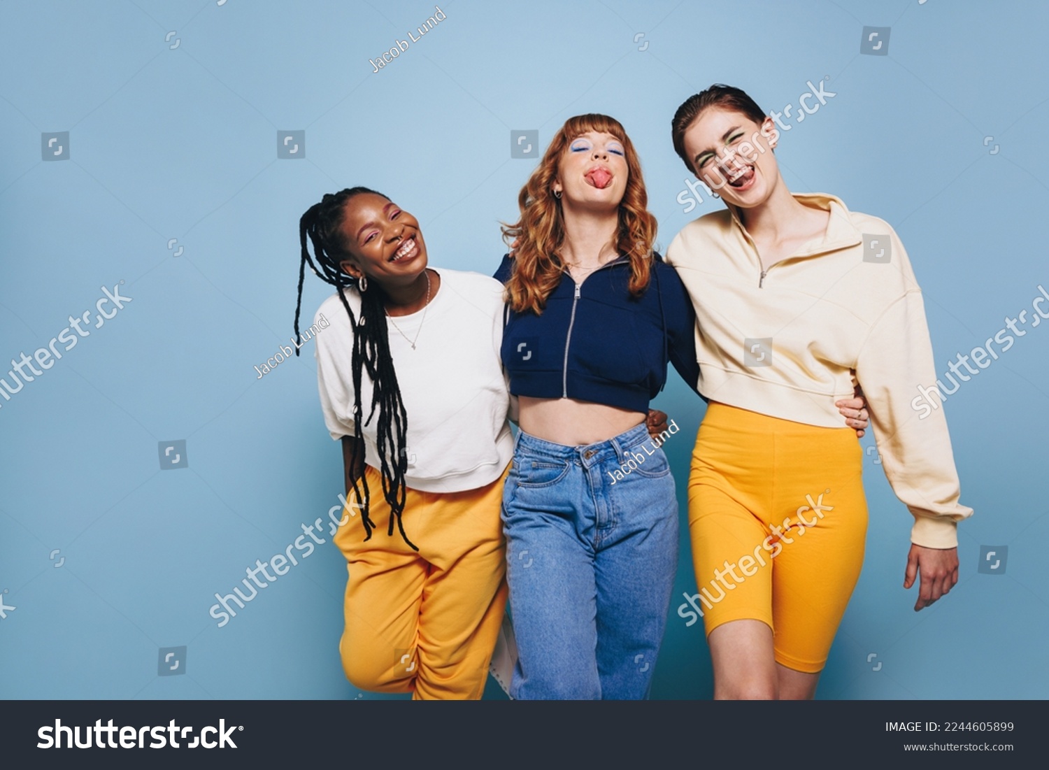 Cheerful female friends making funny faces and looking at the camera while embracing each other. Group of happy young women having fun while standing against a blue studio background. #2244605899