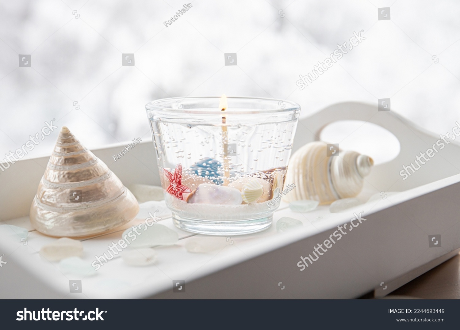 Homemade underwater seashell clear gel wax candle inside clear cup. Beautiful candle on white wood tray with sea glass and seashells on window sill at home. #2244693449