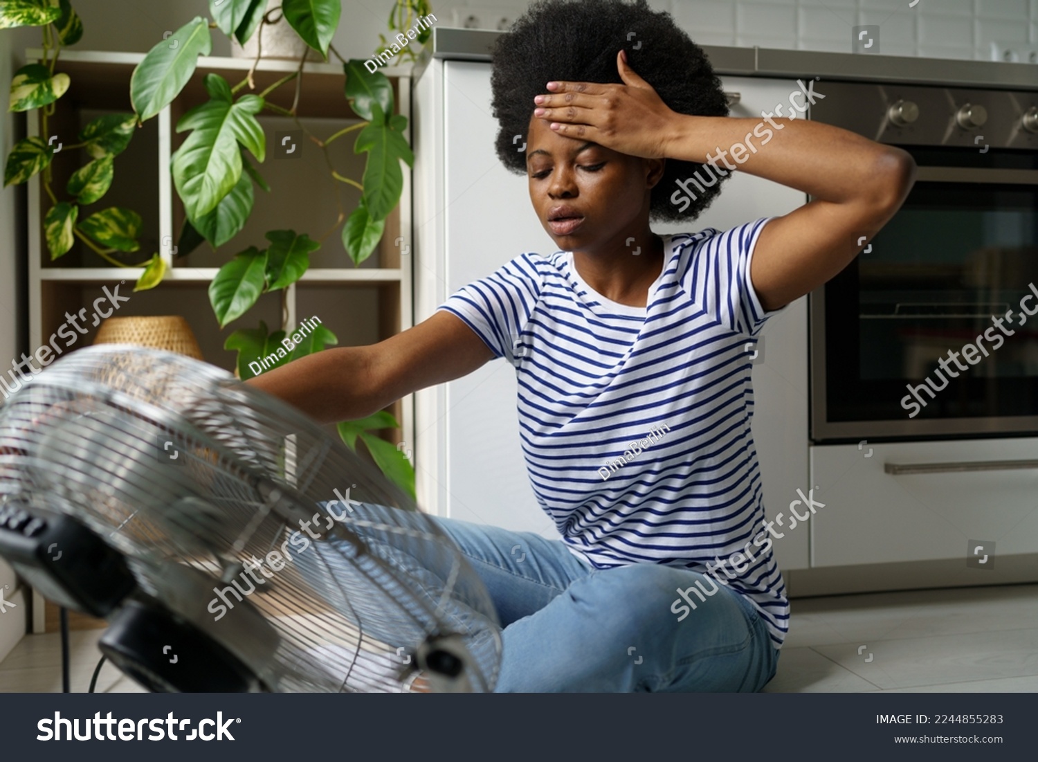 Unhappy African woman touching forehead having hot flashes using electric fan to cool off, overheated black female sitting in front of air cooler at home, surviving heatwave without air conditioning #2244855283