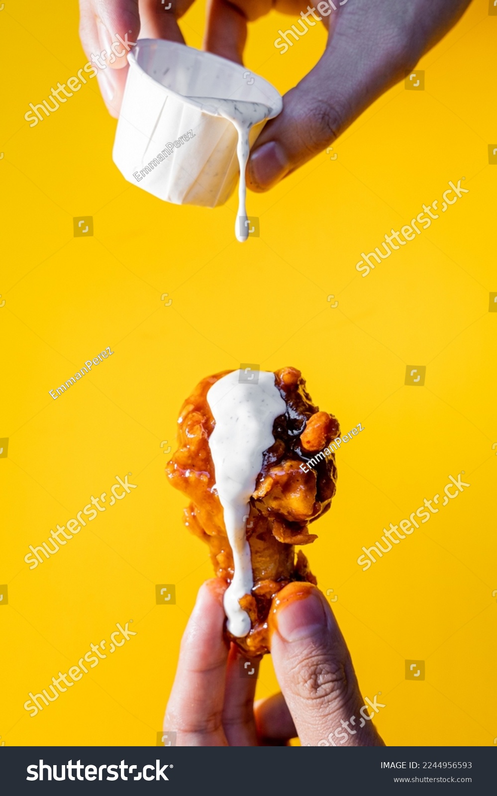 Hand holding a chicken wing with buffalo and ranch dressing on a yellow background. #2244956593