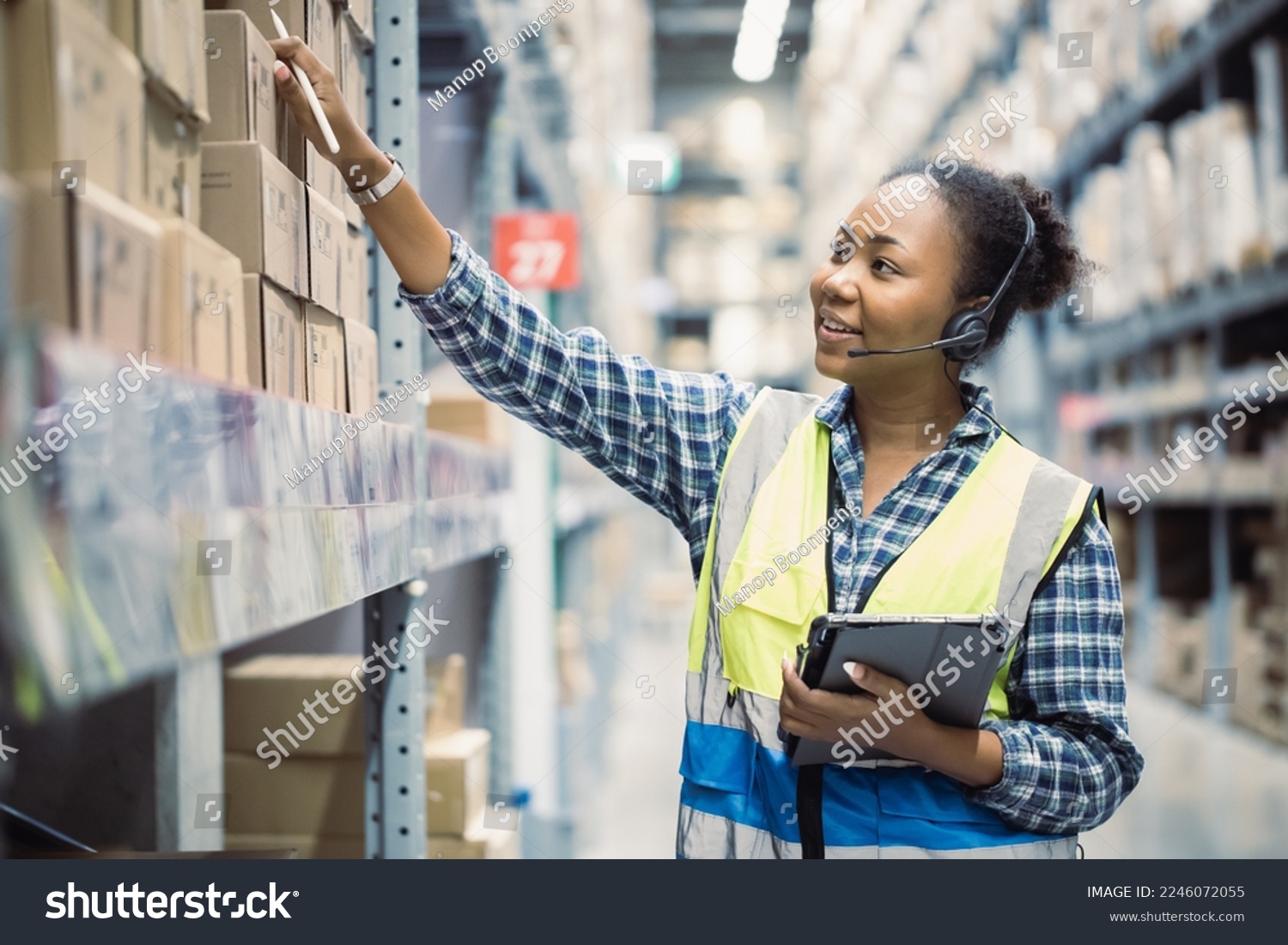 Portrait of young attractive African American woman auditor or trainee staff work looking up stocktaking inventory in warehouse store by computer tablet and headphones near products shelf. Call center #2246072055