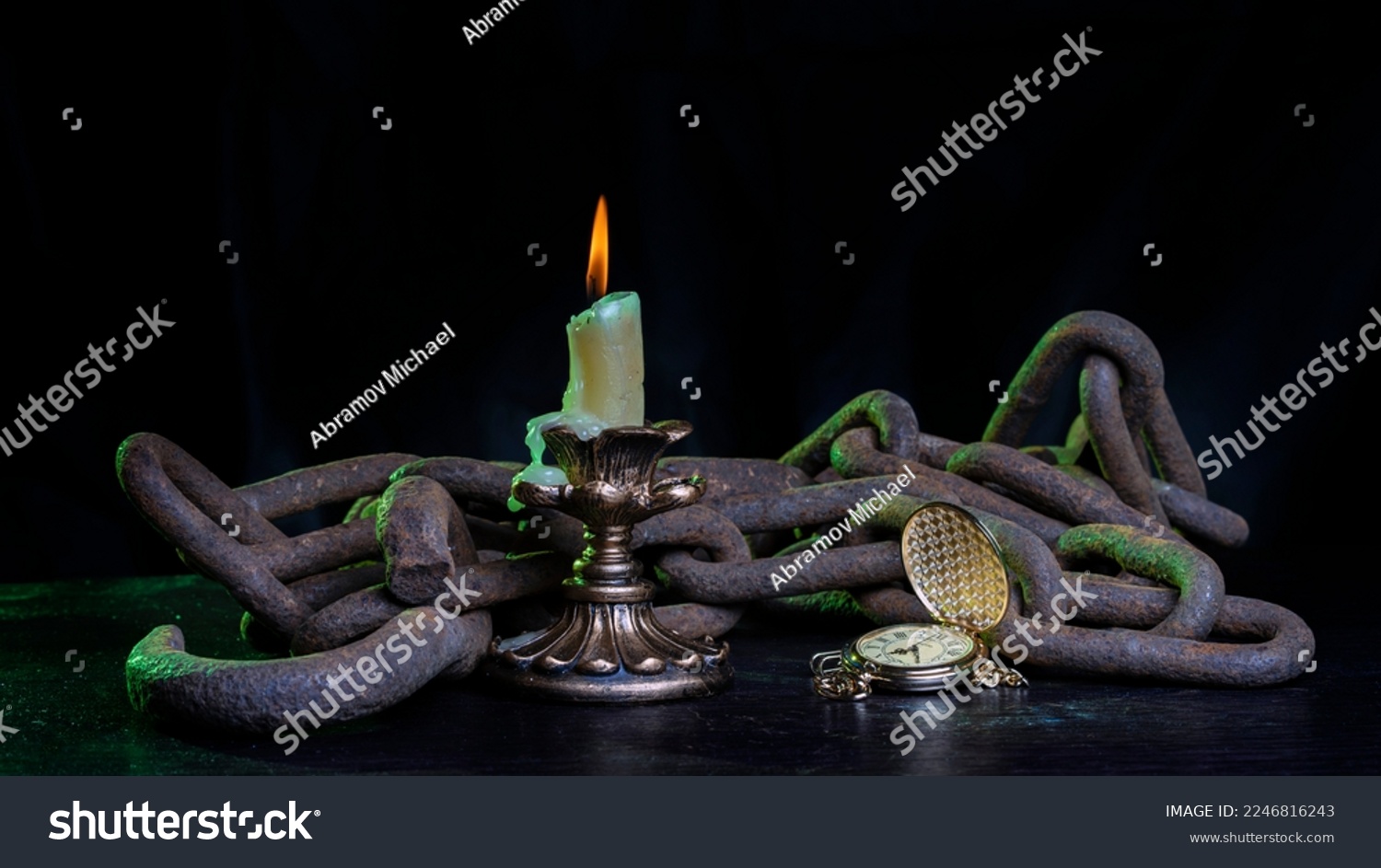 A burning candle in chains on a wooden table and a clock that has stopped time #2246816243