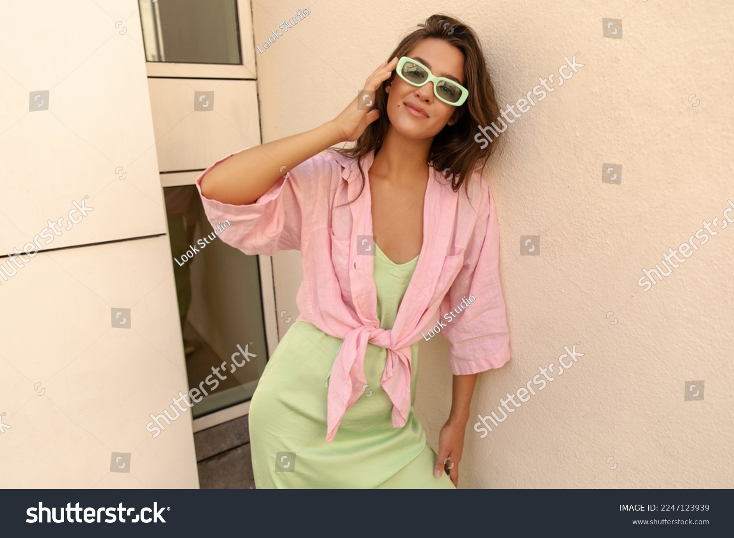 Fashionable young caucasian woman looking at camera corrects her sunglasses. Model with brunette hair wears pink shirt, sundress and sunglasses. Great mood lifestyle concept. #2247123939