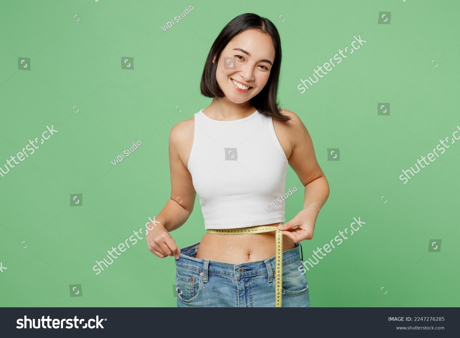 Young woman wear white clothes show loose pants after weightloss hold measure tape on waist isolated on plain pastel light green background. Proper nutrition healthy fast food unhealthy choice concept #2247276285