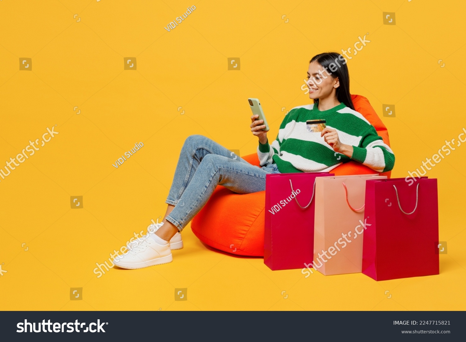 Full body happy young latin woman wear green knitted sweater sit in bag chair near package bags after shopping use mobile cell phone isolated on plain yellow background Black Friday sale buy concept #2247715821