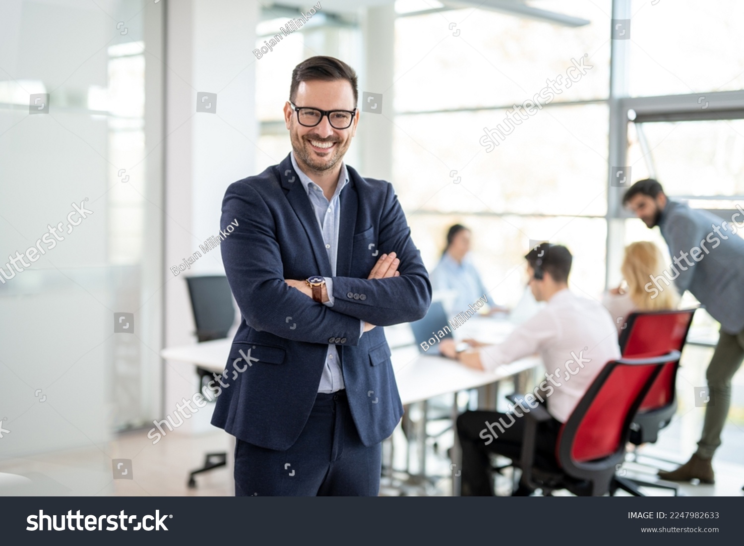 Portrait of confidence business man in formalwear standing with crossed hands at office with colleagues in the background #2247982633